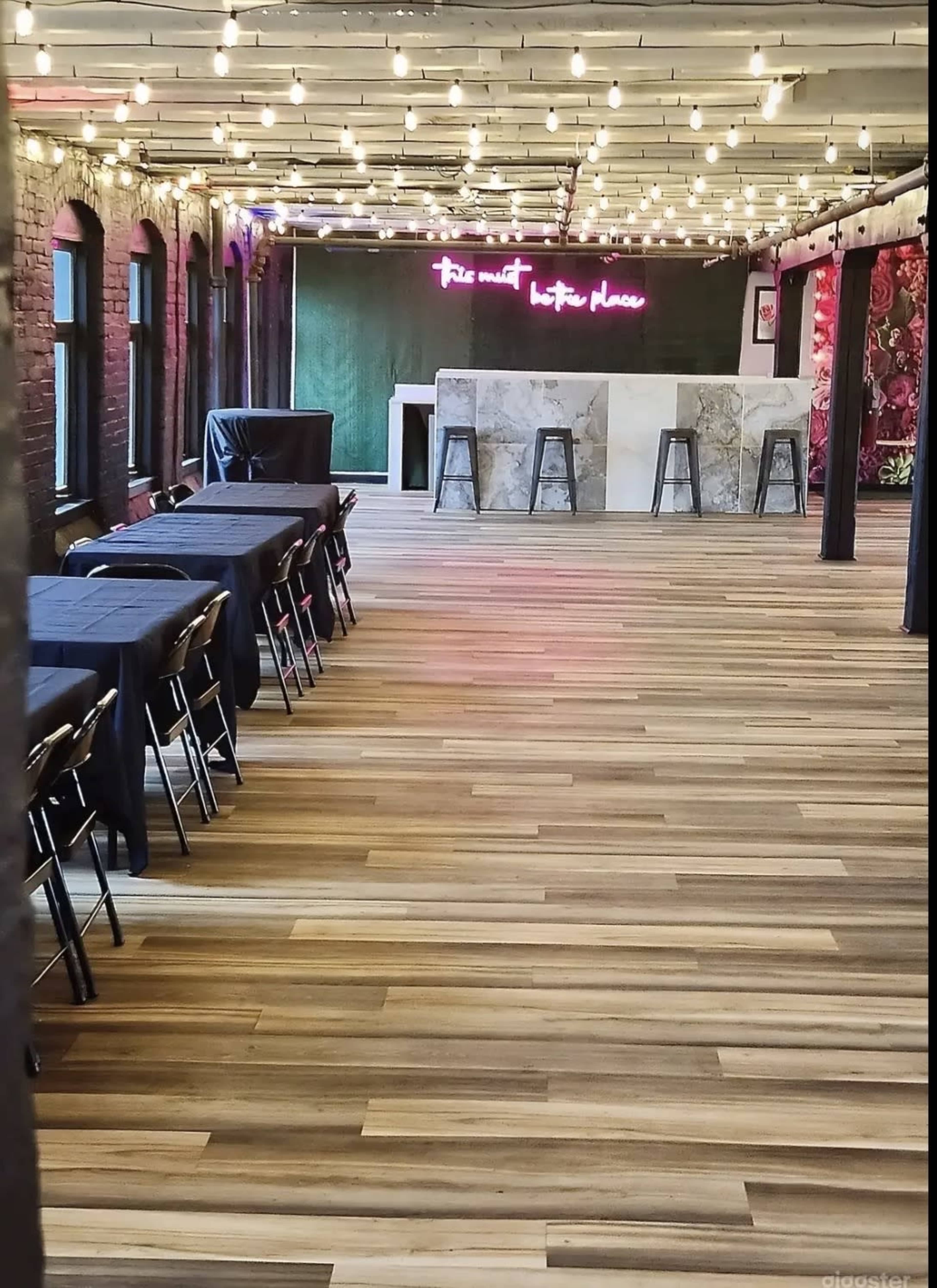 The image shows an empty venue with wooden flooring, tables covered in black cloths, and string lights overhead, along with a bar area featuring a neon sign.
