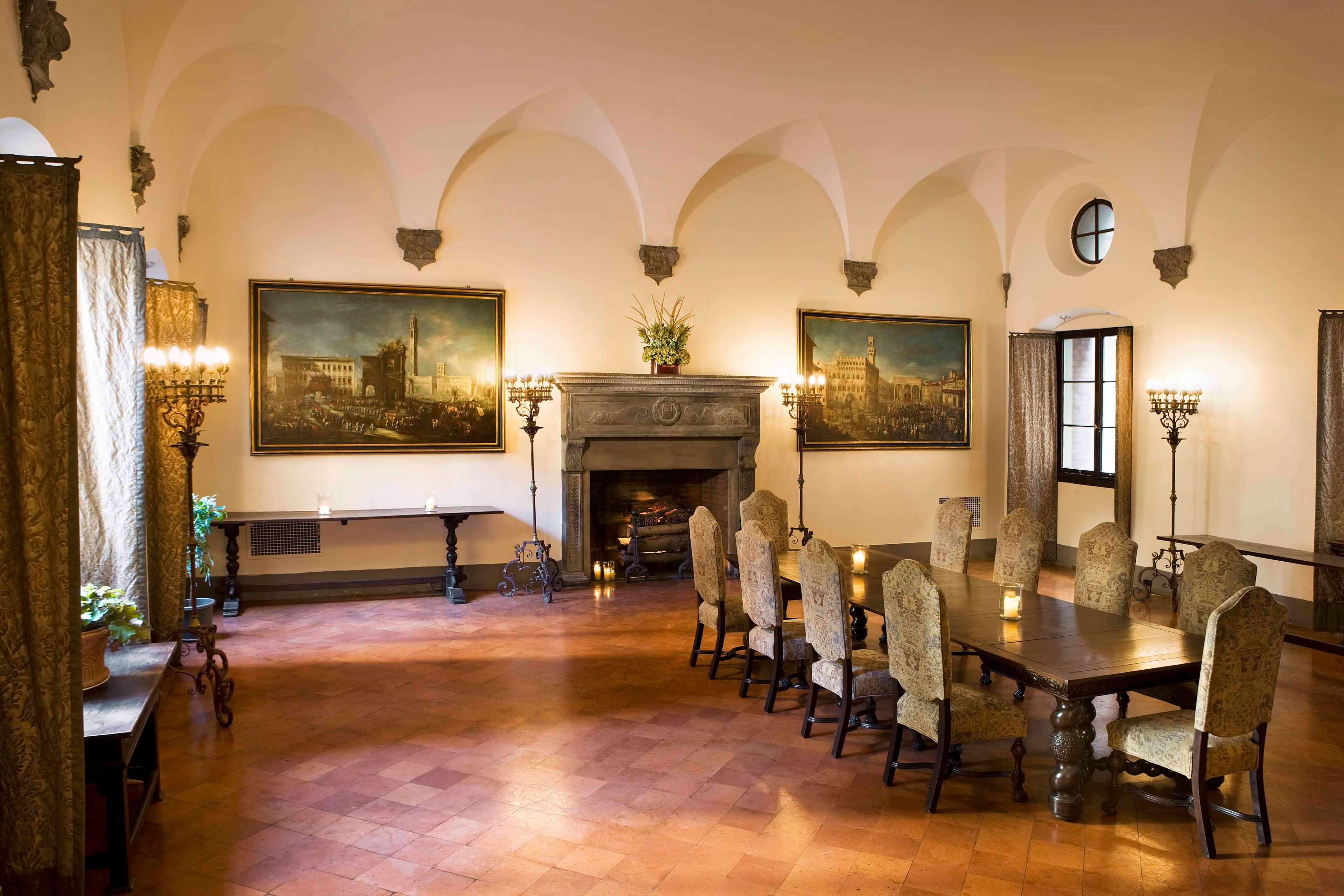 The image depicts a spacious dining room with a long wooden table surrounded by upholstered chairs, flanked by large paintings and a stone fireplace.