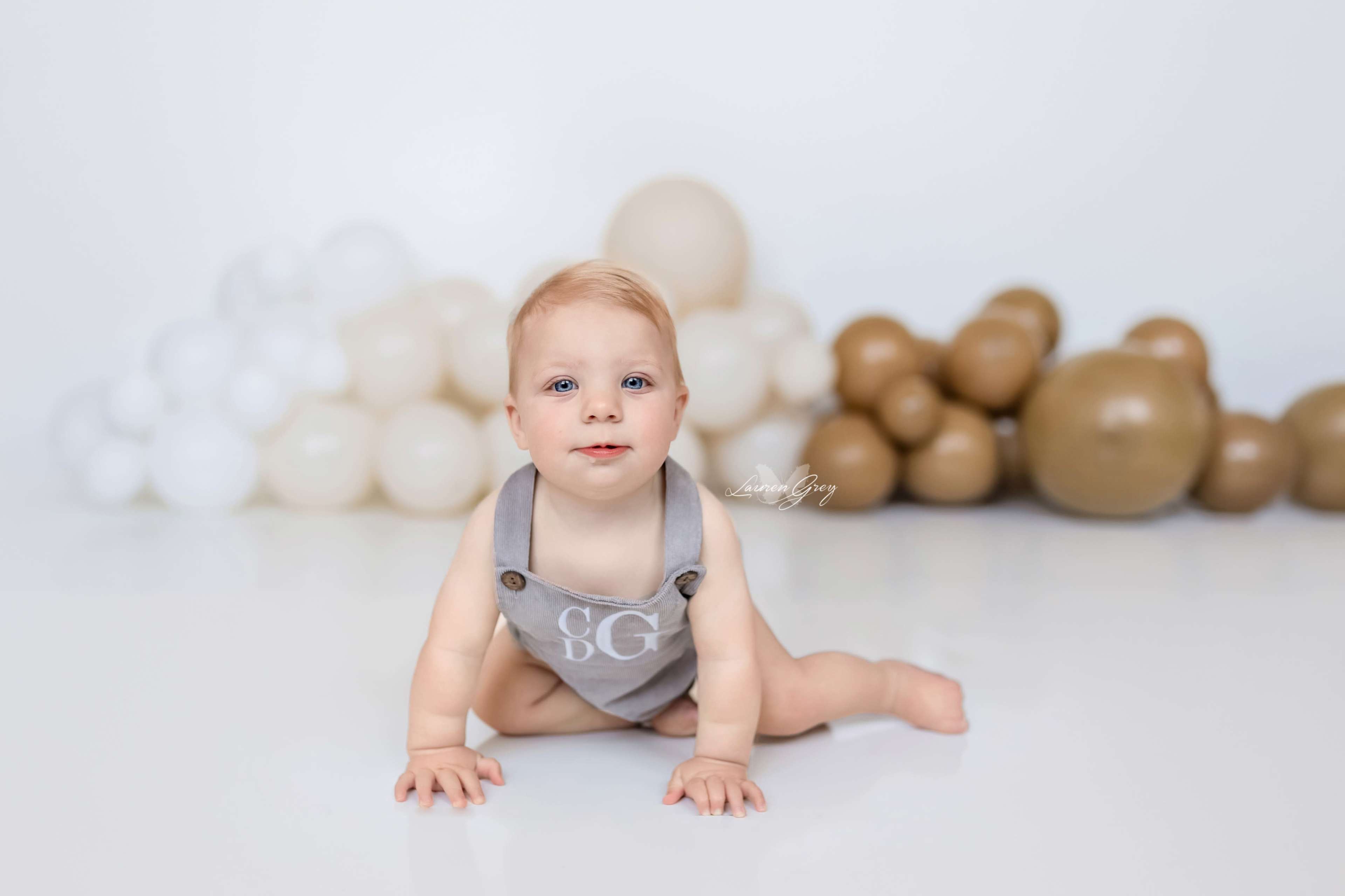 A baby in a gray outfit with the initials "SG" crawls on a white floor in front of a backdrop of beige and brown balloons.