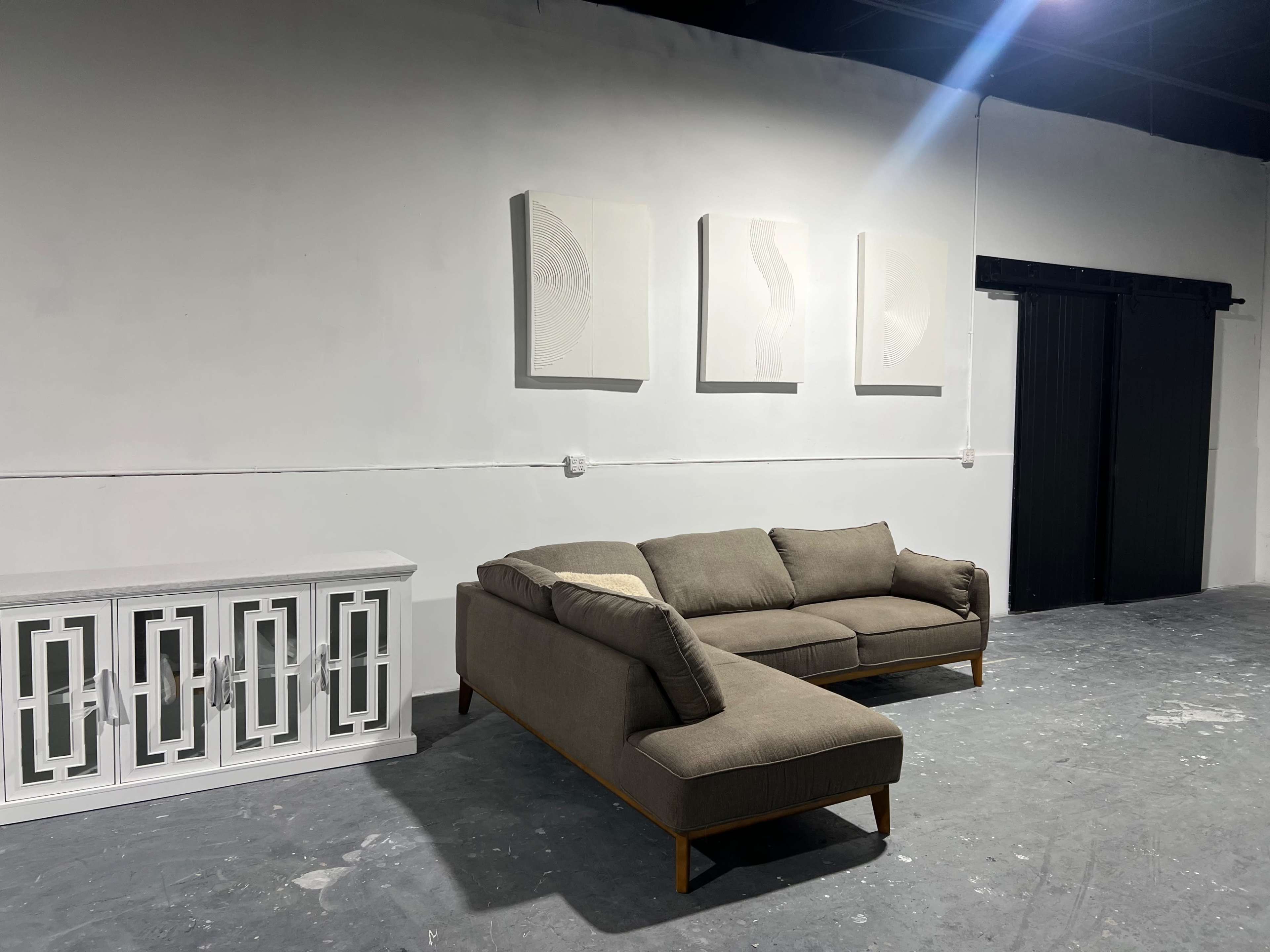 A gray sectional couch sits against a white wall, accompanied by three abstract white artworks above it and a white sideboard nearby.