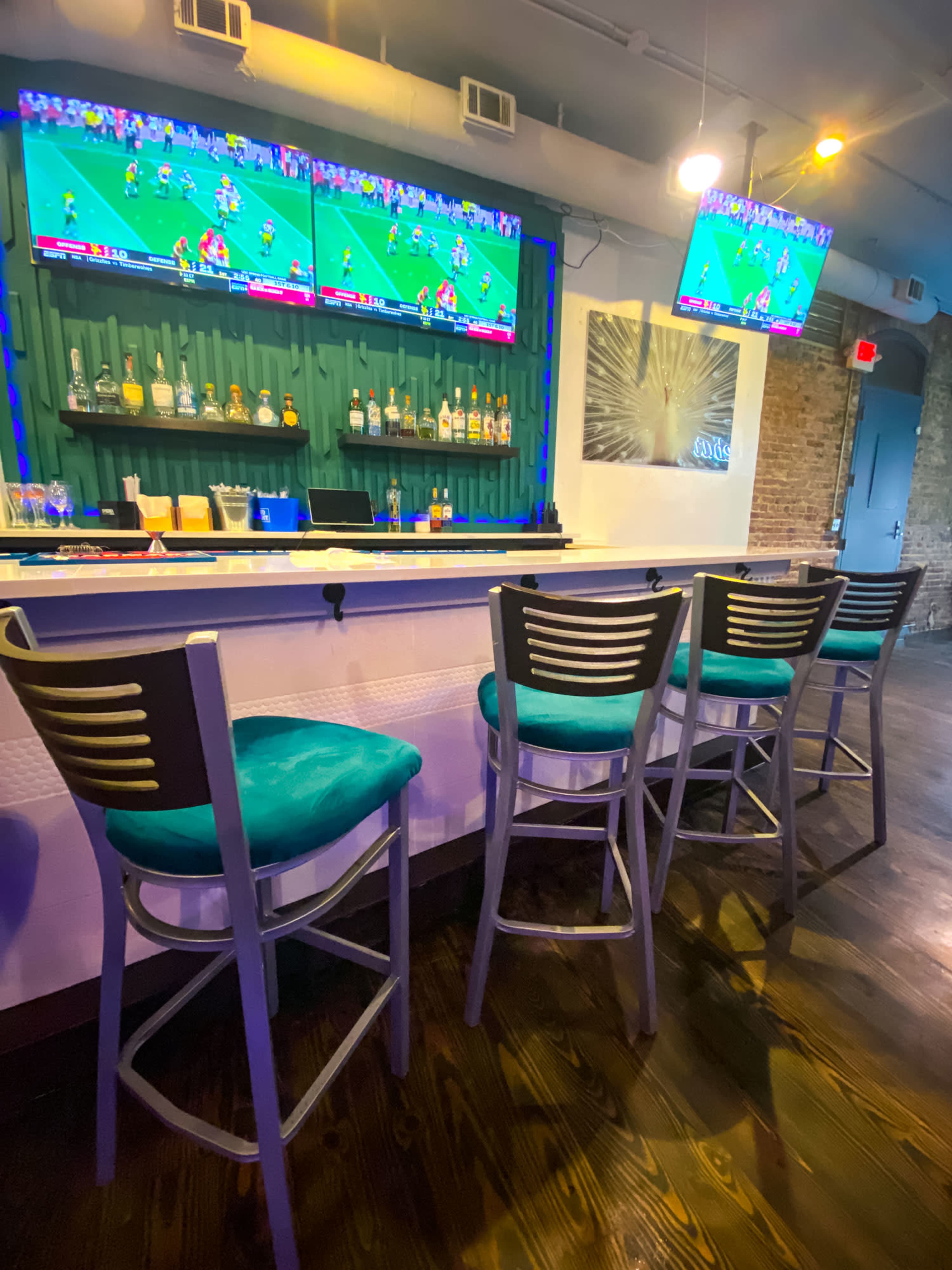 The image shows a bar area with three empty stools and two televisions displaying a football game on the wall behind a backlit shelf of liquor bottles.