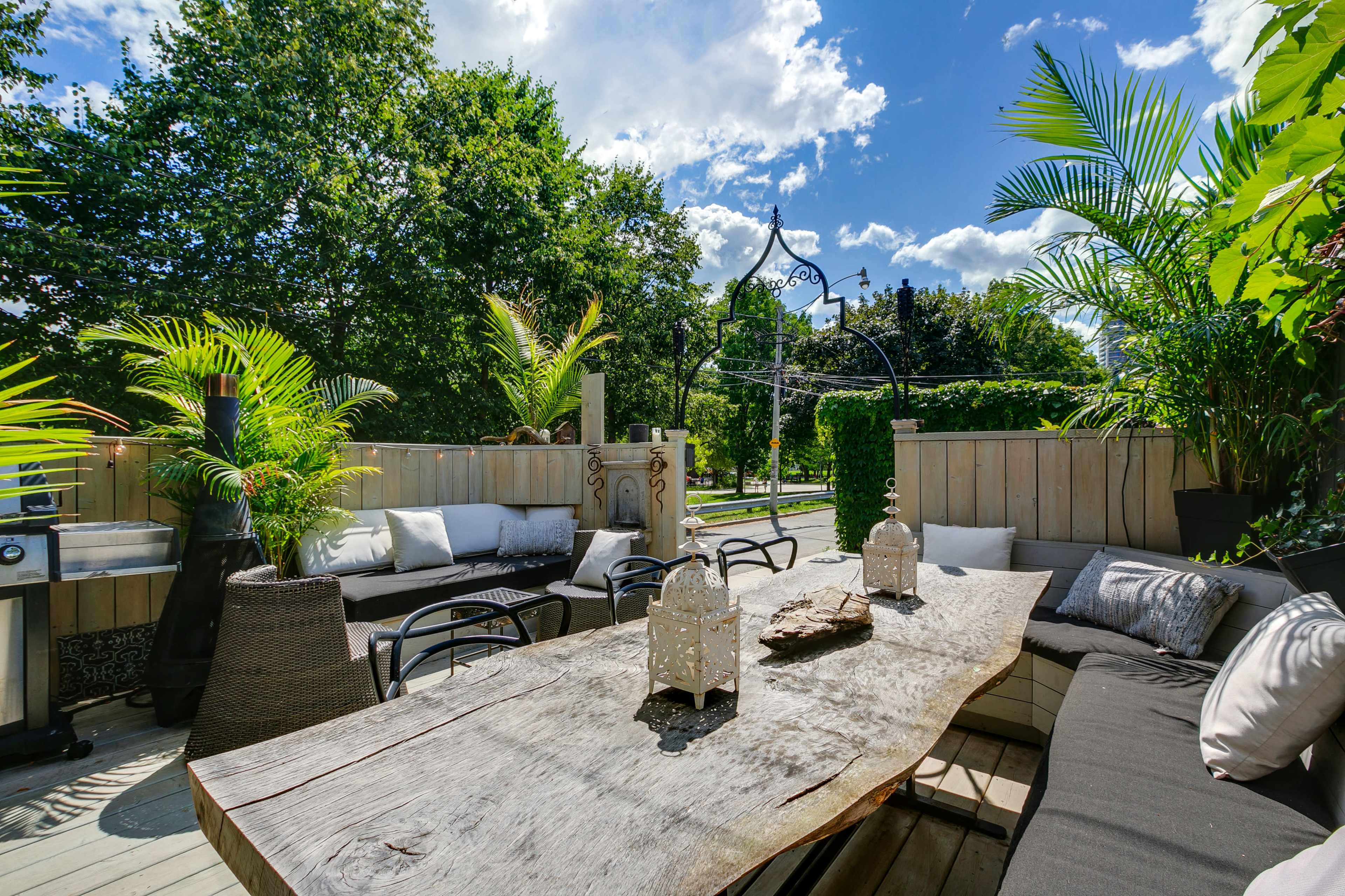 A wooden dining table is surrounded by outdoor seating, with potted plants and decorative lanterns set against a backdrop of trees and a bright sky.