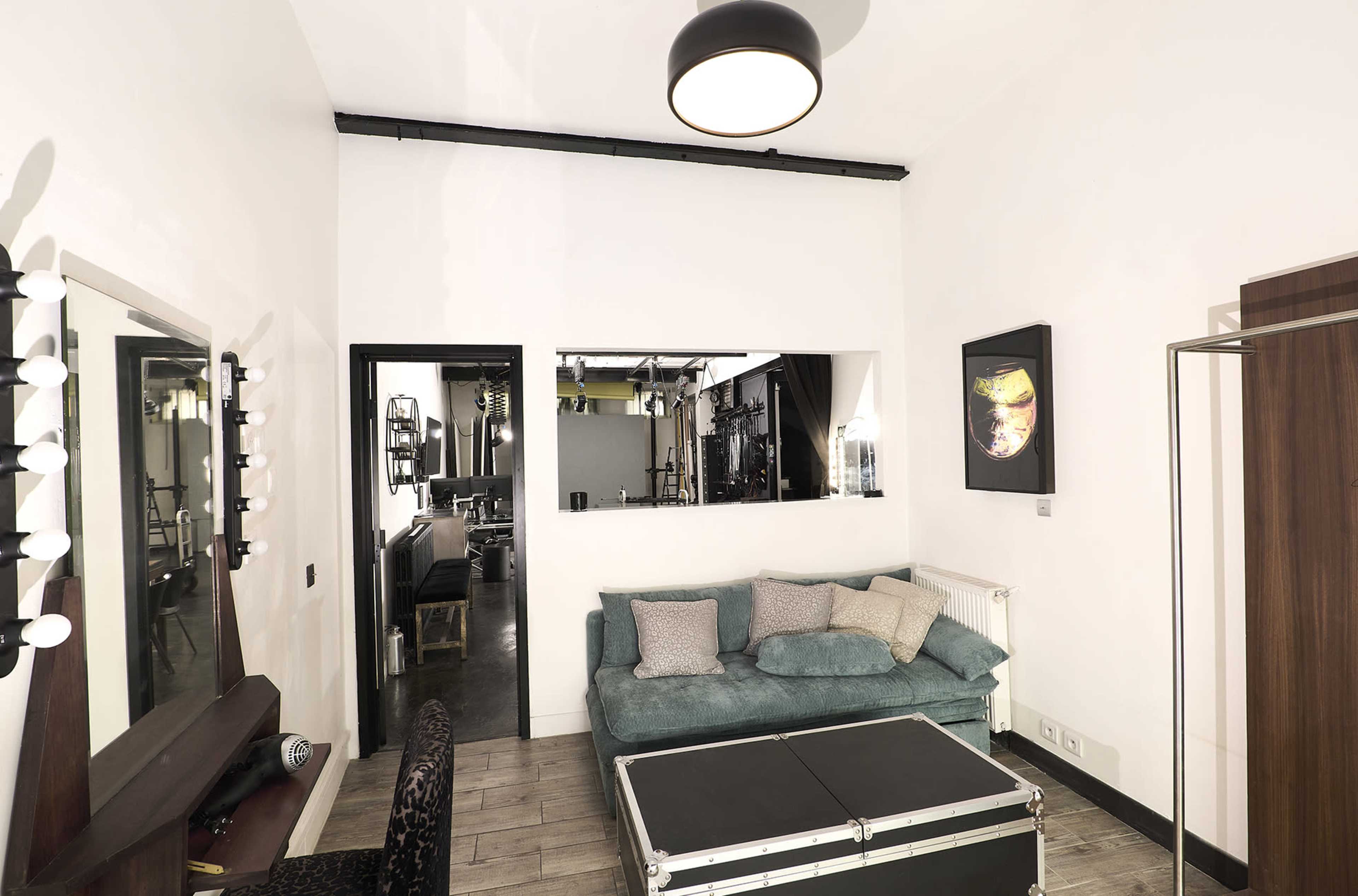 The image shows a small room with a teal couch, a mirror, a dressing table with lights, and a black-framed doorway leading to another area.