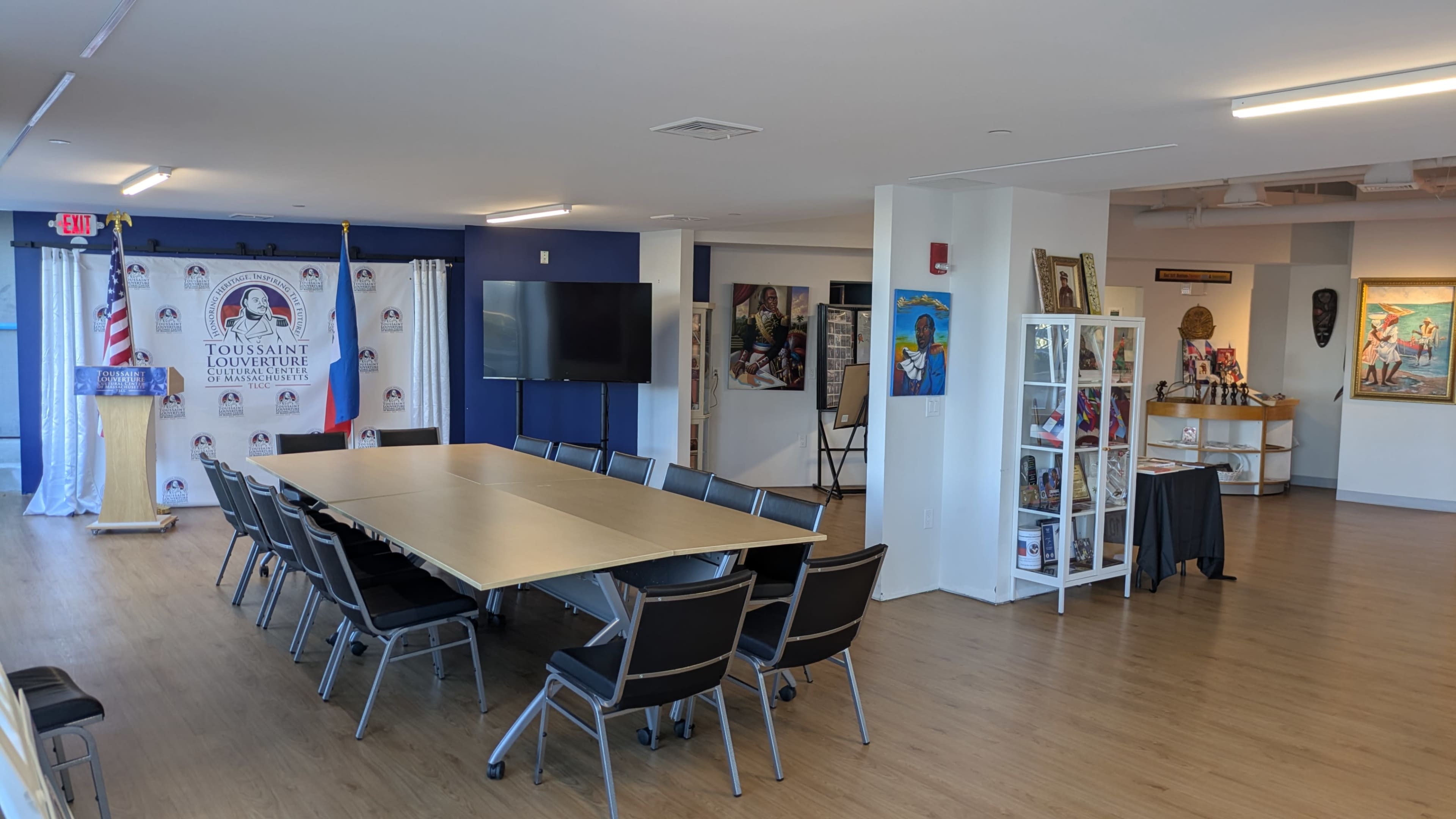 The image shows a meeting room with a long table and chairs, a display area with glass cabinets, artwork on the walls, and a banner featuring the name "Toussaint Louverture" in the background.