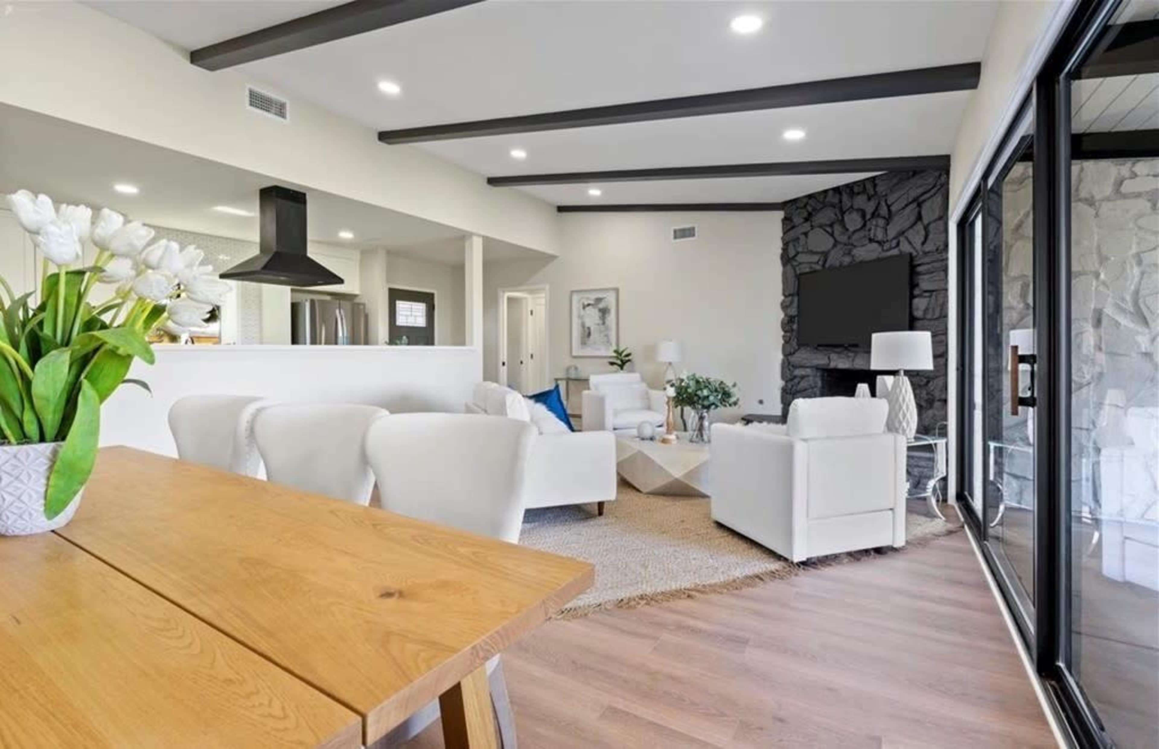 The image shows a light-filled living room with white furniture, a wooden dining table, and a stone fireplace.