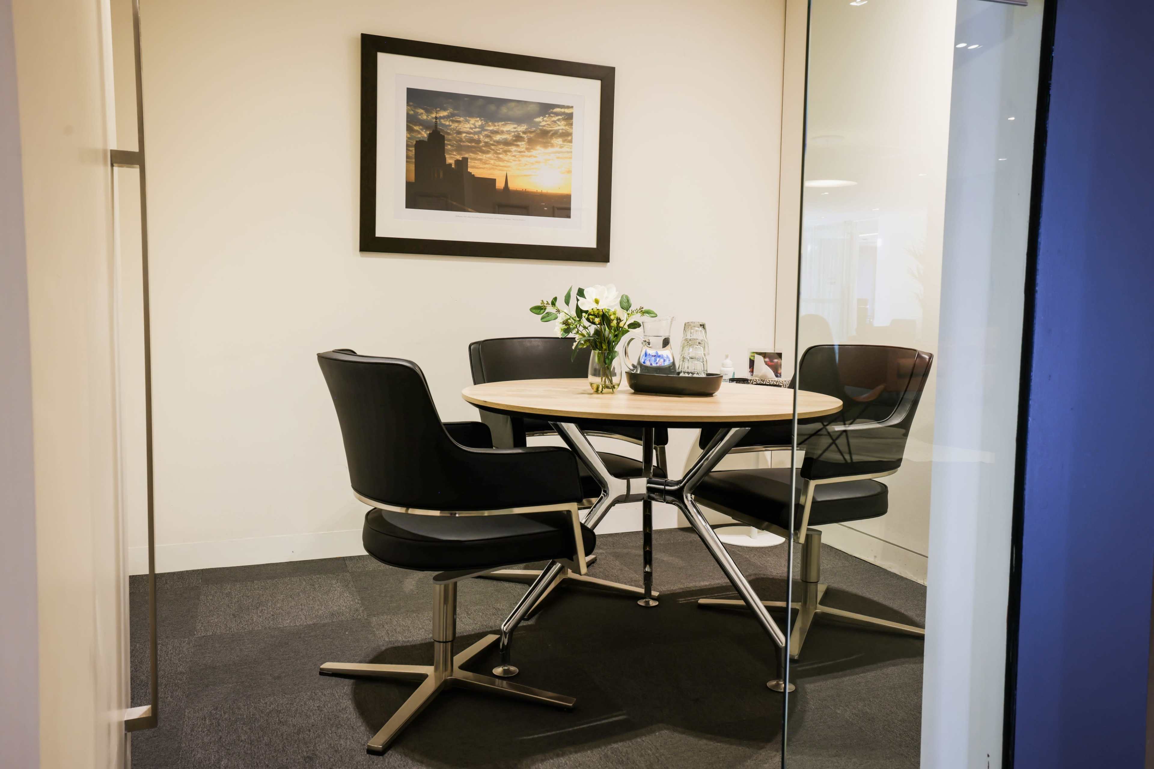 A small meeting room features a round wooden table surrounded by four black chairs, with a framed picture of a sunset on the wall.