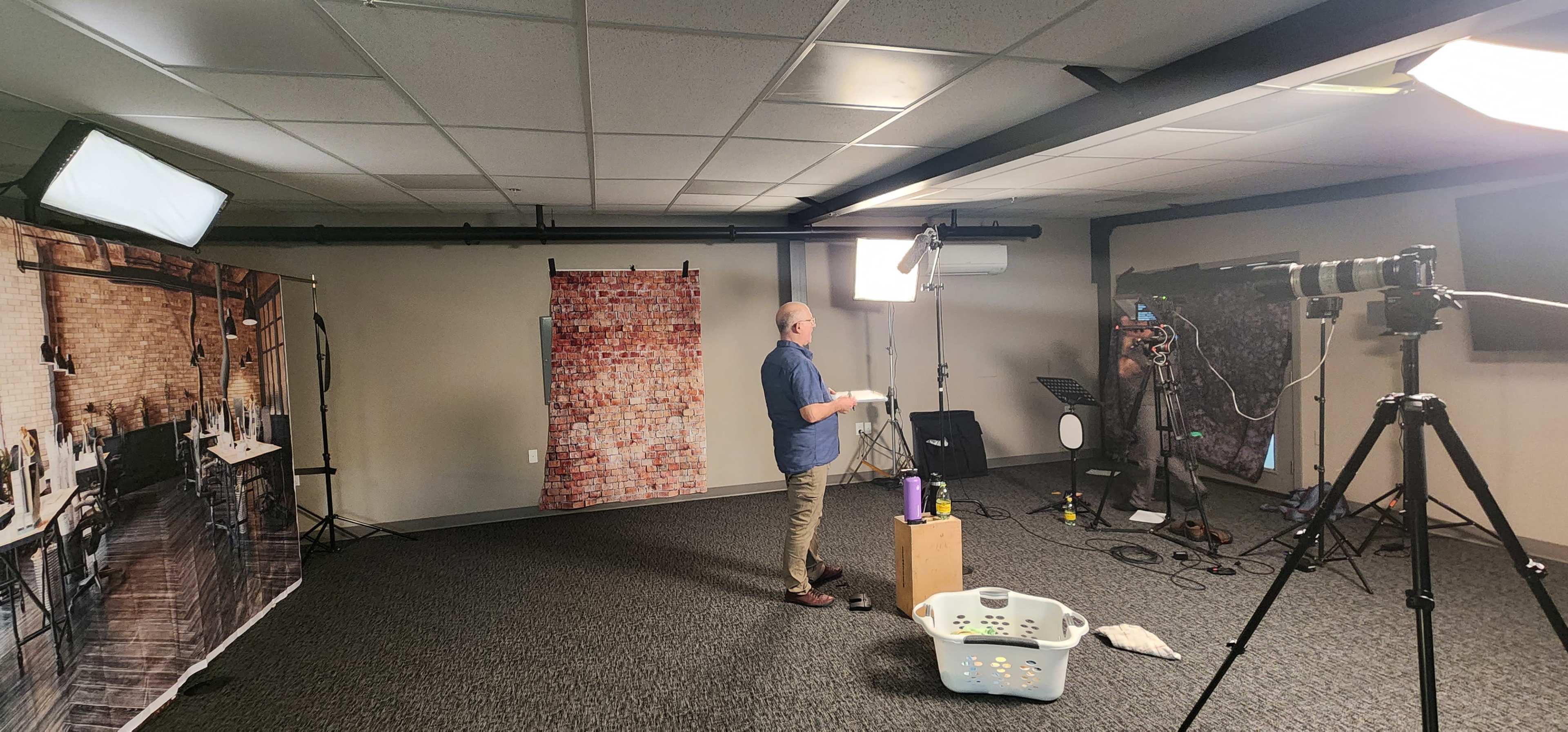 A man stands in a studio setting with various lighting equipment and backdrops while holding a clipboard.