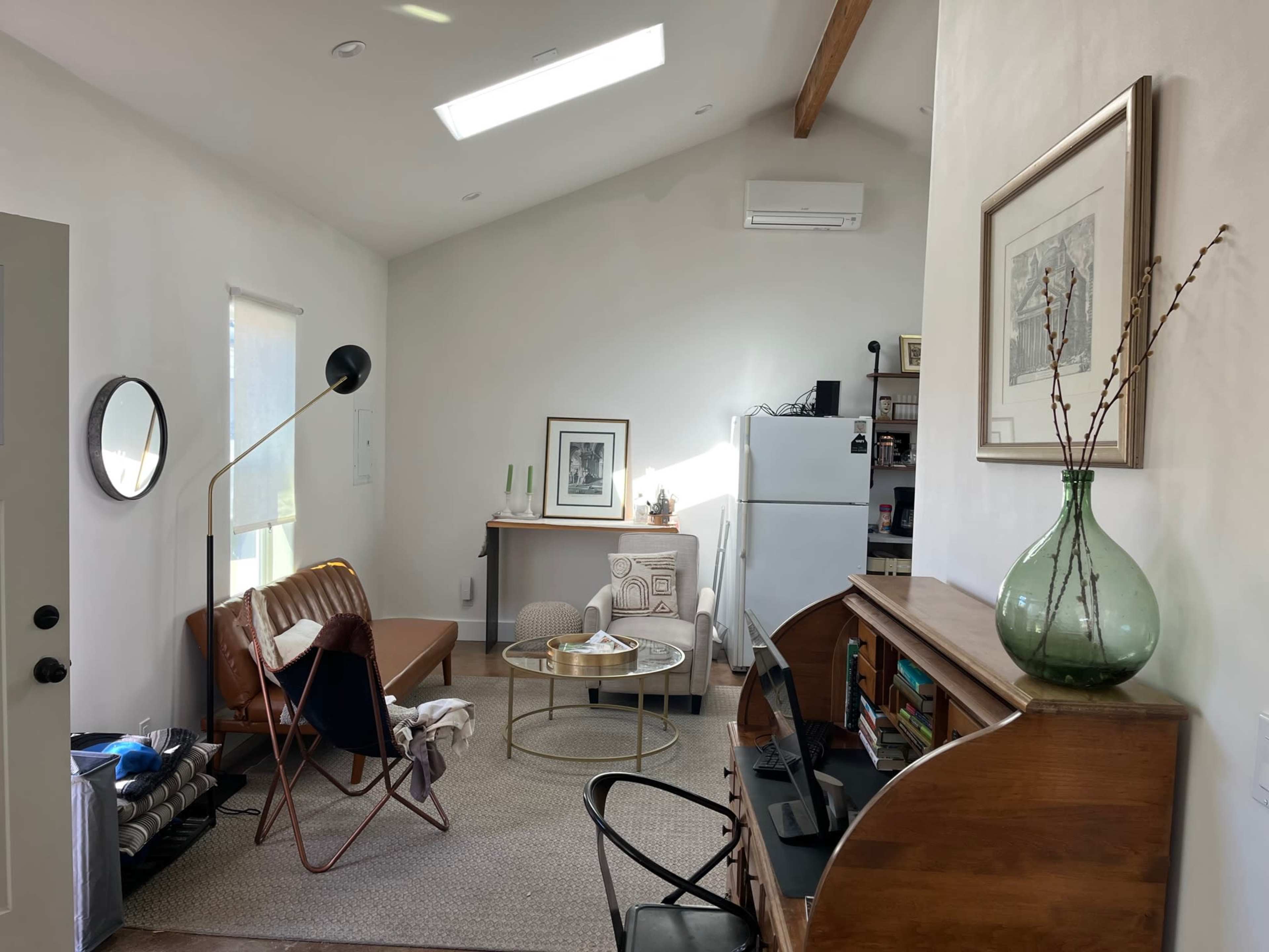 A modern living room with a sofa, a round coffee table, an armchair, and a wooden desk, featuring a large window and a refrigerator.