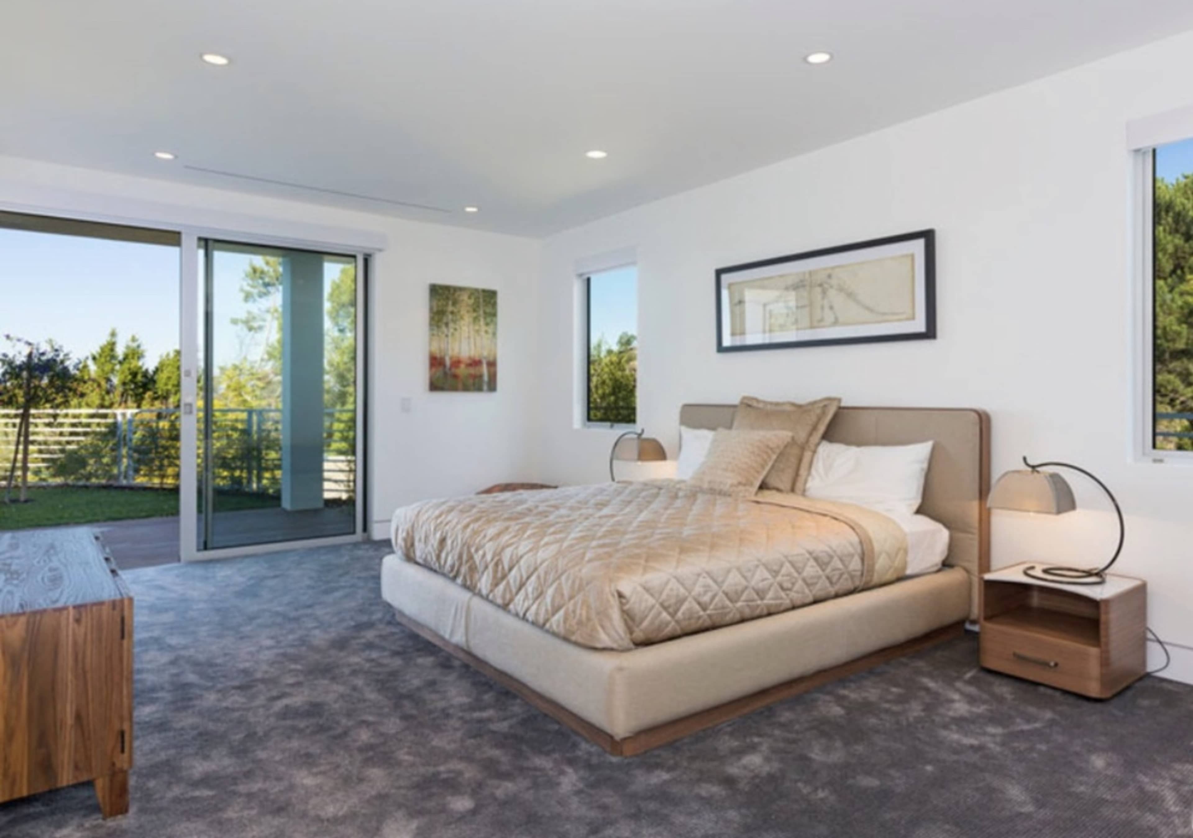 A modern bedroom featuring a large bed with a patterned comforter, two bedside lamps, a wooden dresser, and large windows leading to a balcony with a view of greenery.