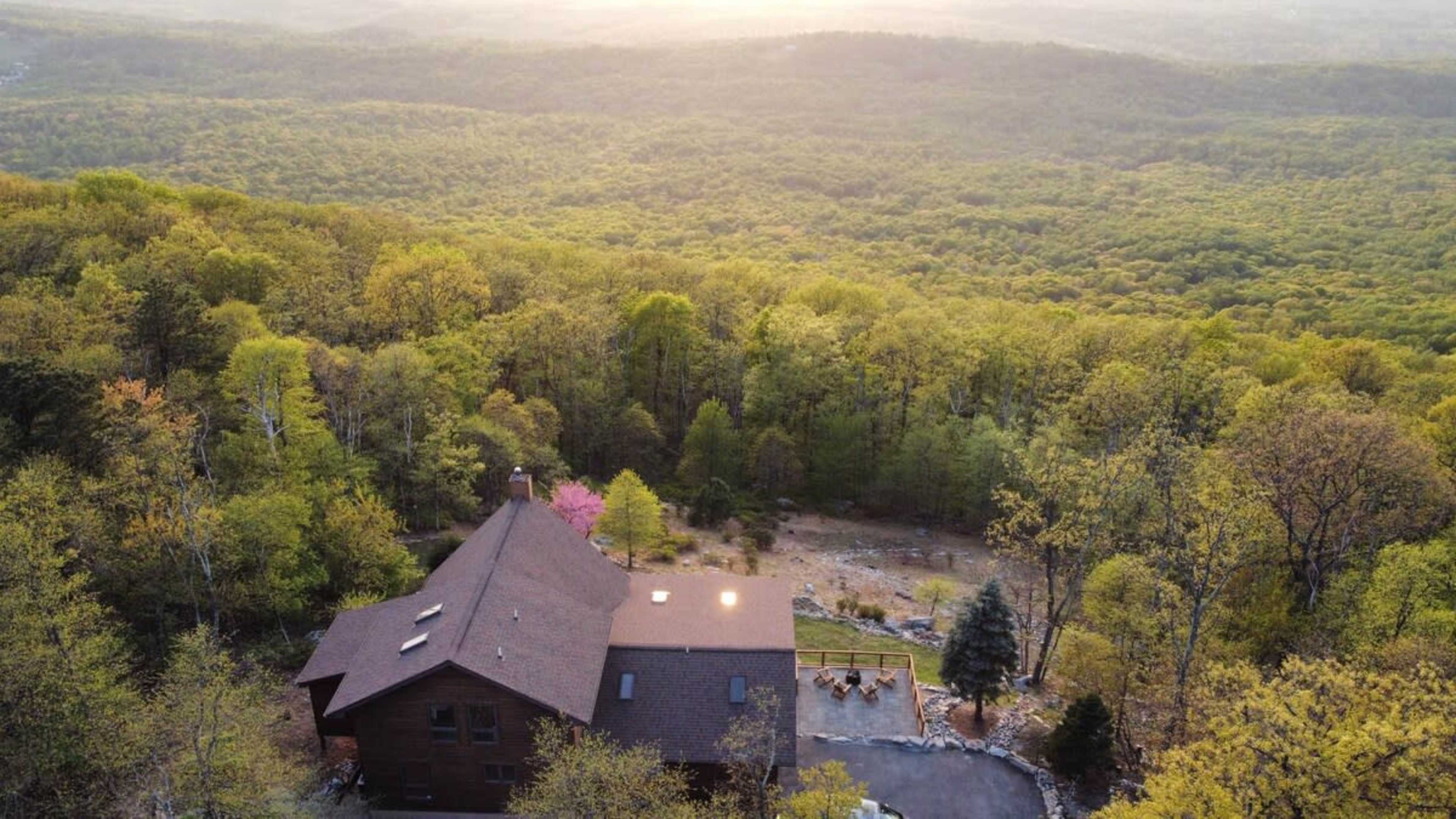 Aerial view of a large wooden house situated on a hillside surrounded by lush green trees and a distant mountainous landscape.