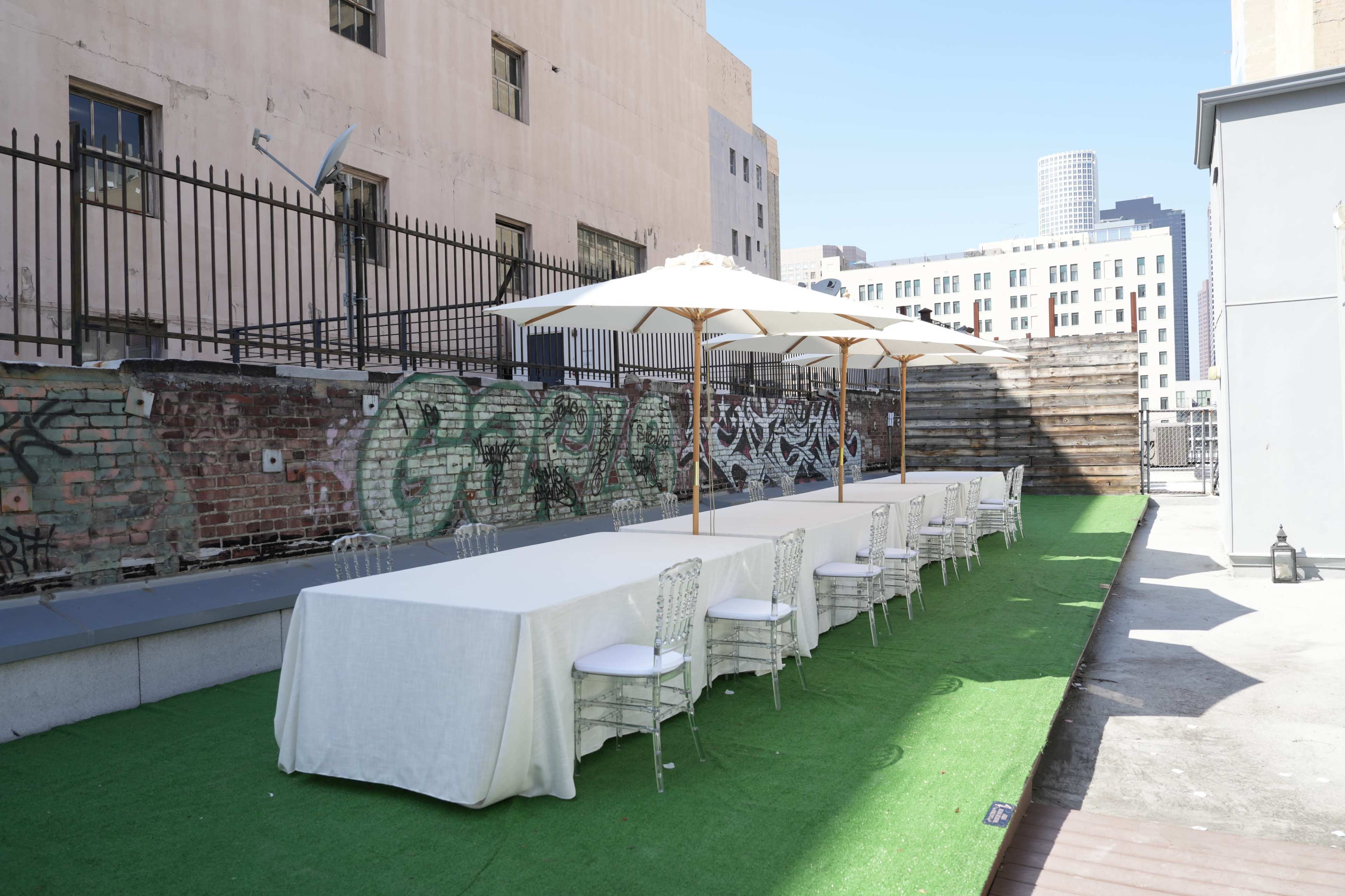 A long, white table is set outdoors on a rooftop, surrounded by decorative umbrellas and green artificial grass, with a backdrop of urban buildings and graffiti.