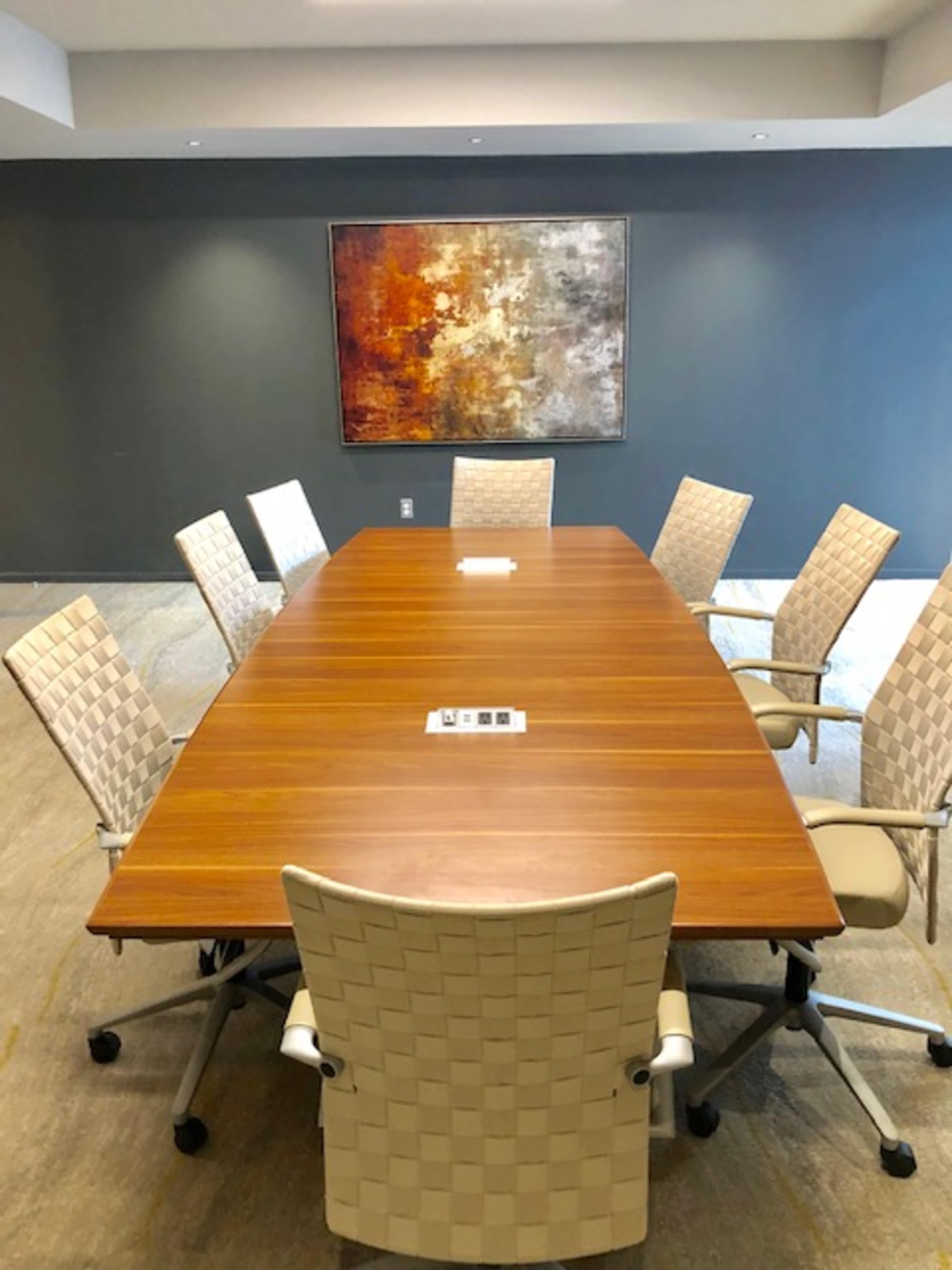 A large wooden conference table surrounded by six modern chairs is set in a room with a gray wall and a colorful abstract painting.