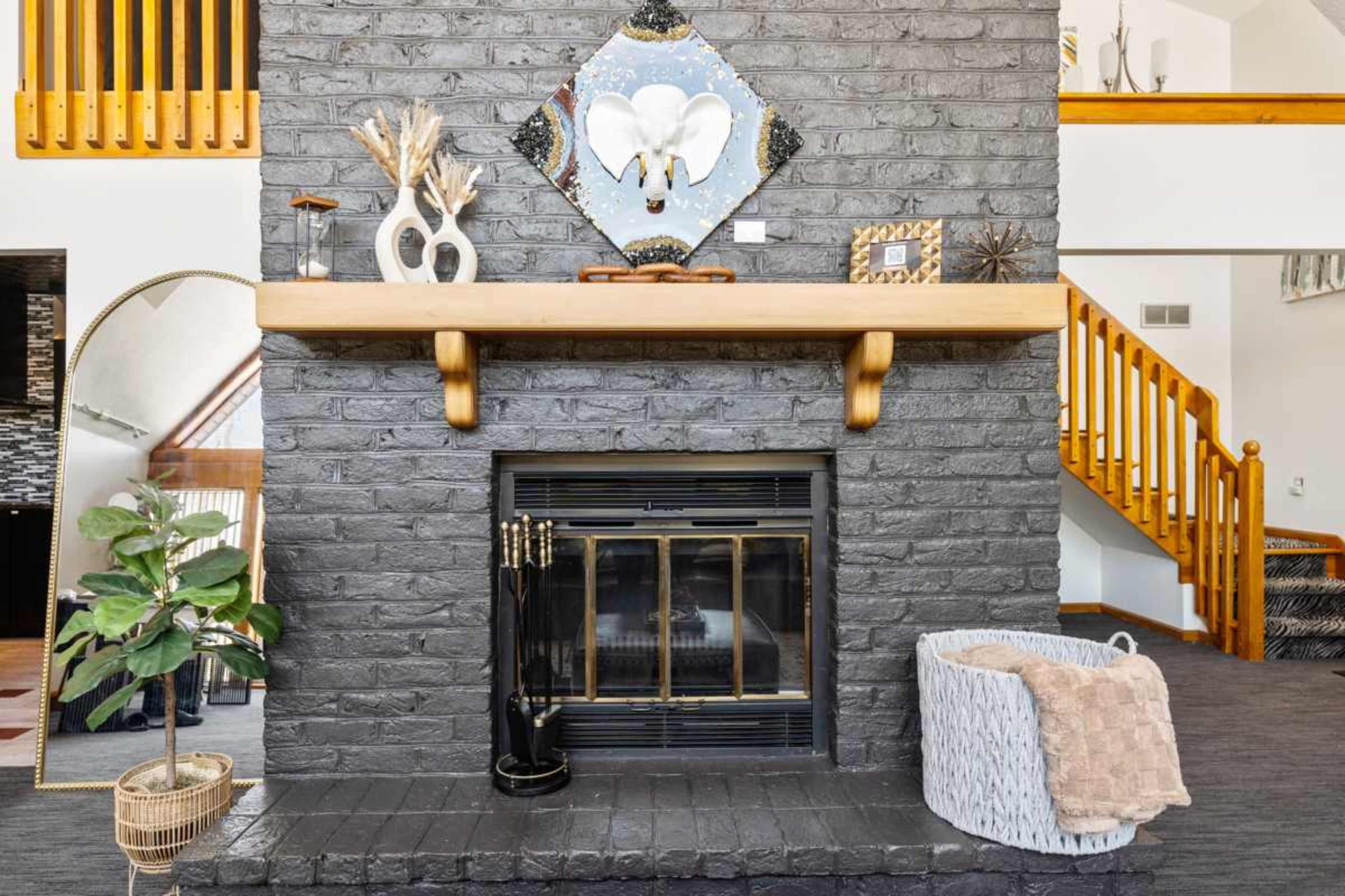 The image shows a stylish living room with a black brick fireplace, a wooden mantel, and decorative items, including a mirror and a potted plant.