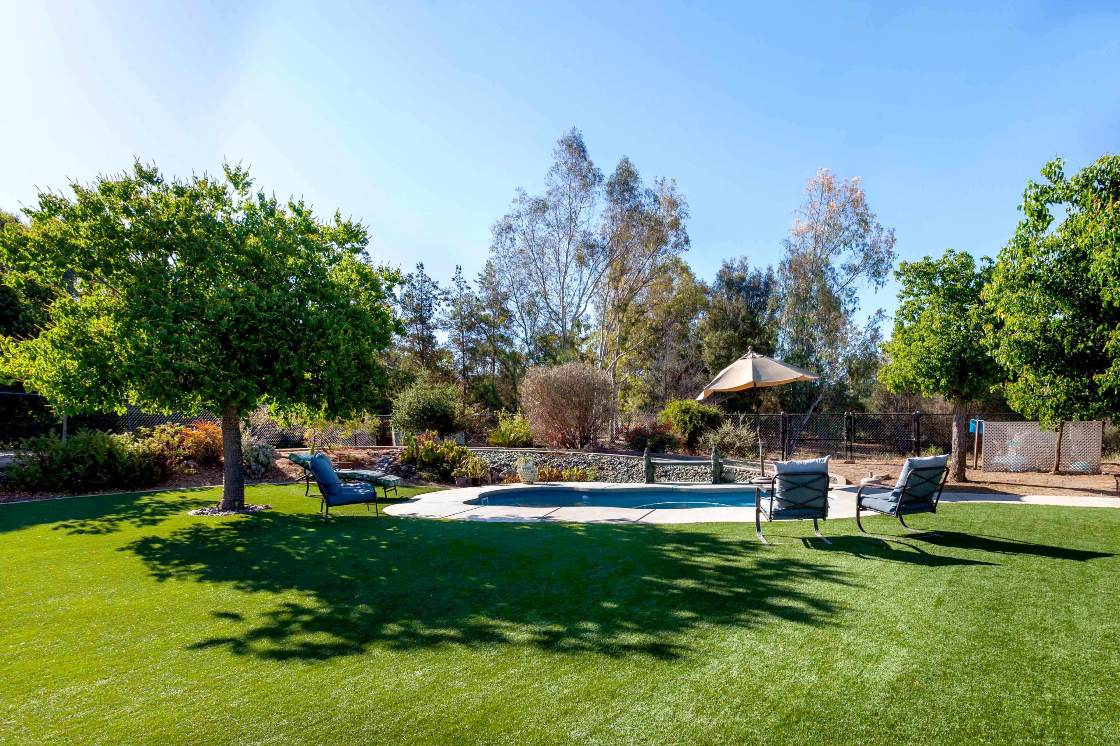 A landscaped backyard with green grass, a small swimming pool, a shaded area under an umbrella, and two lounge chairs.