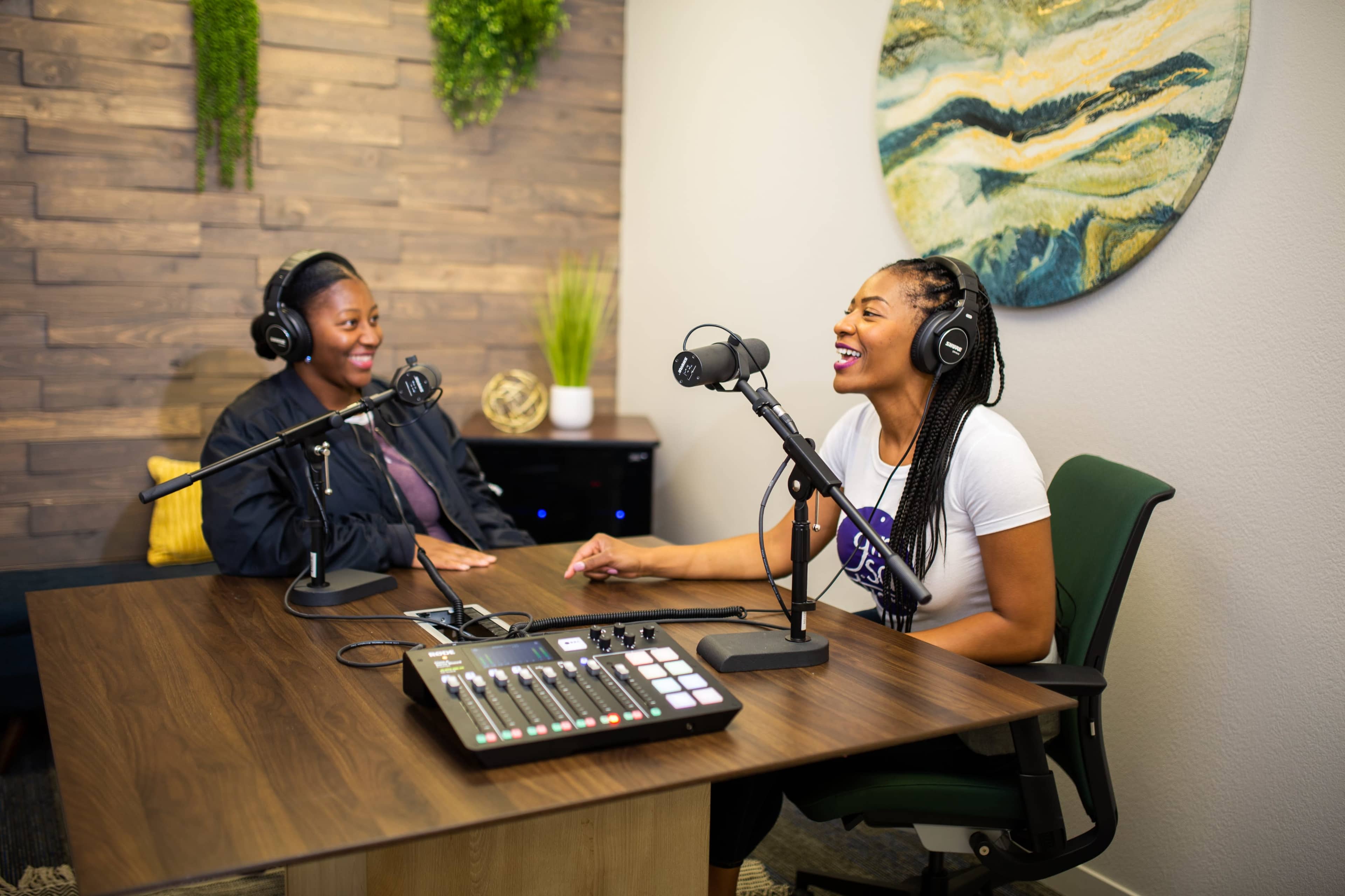 Two women are seated in a podcast studio, engaging in conversation while using microphones and a sound mixing board between them.