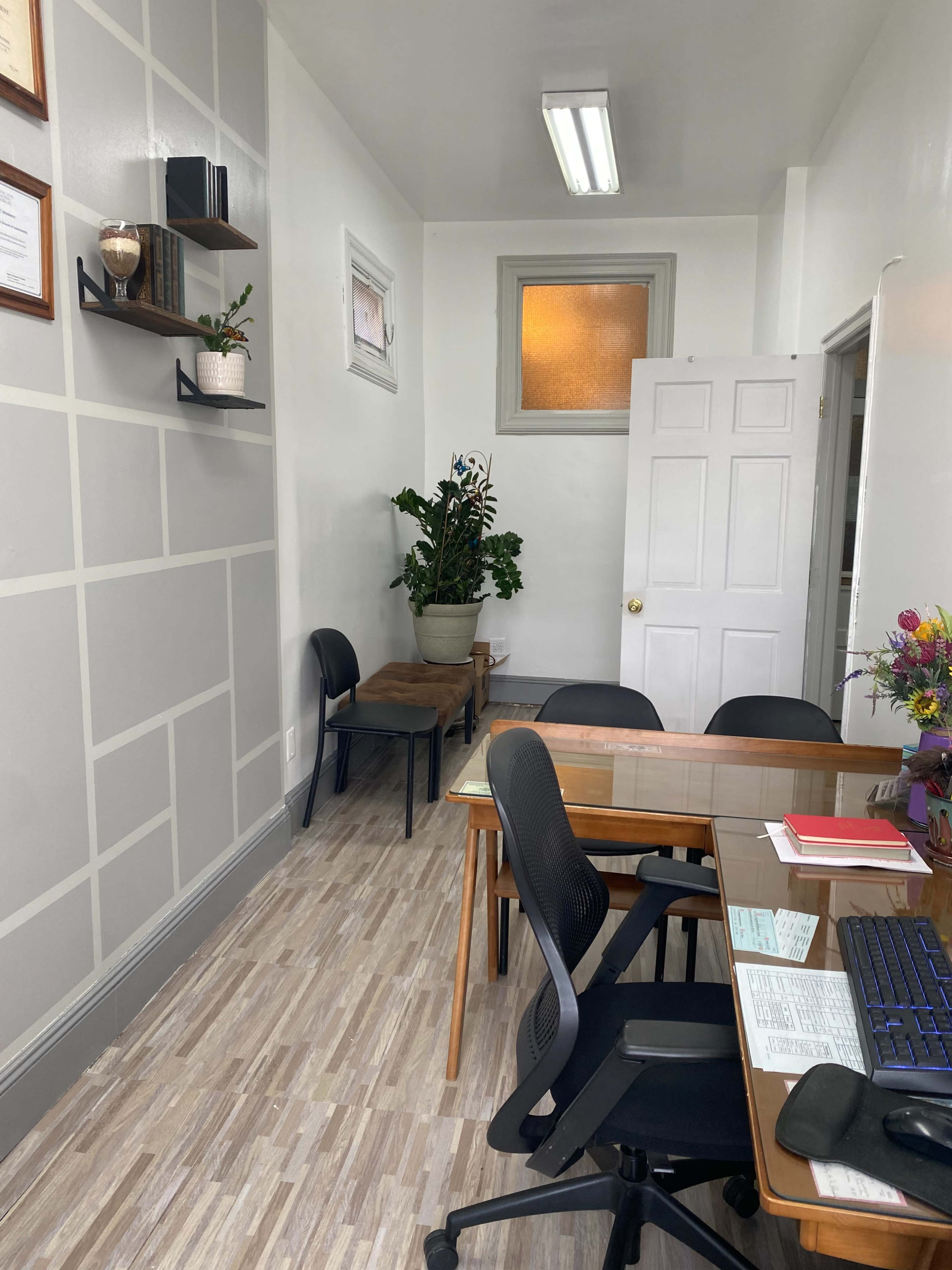 A small, tidy office space with a wooden desk, several black chairs, and a potted plant in the corner.