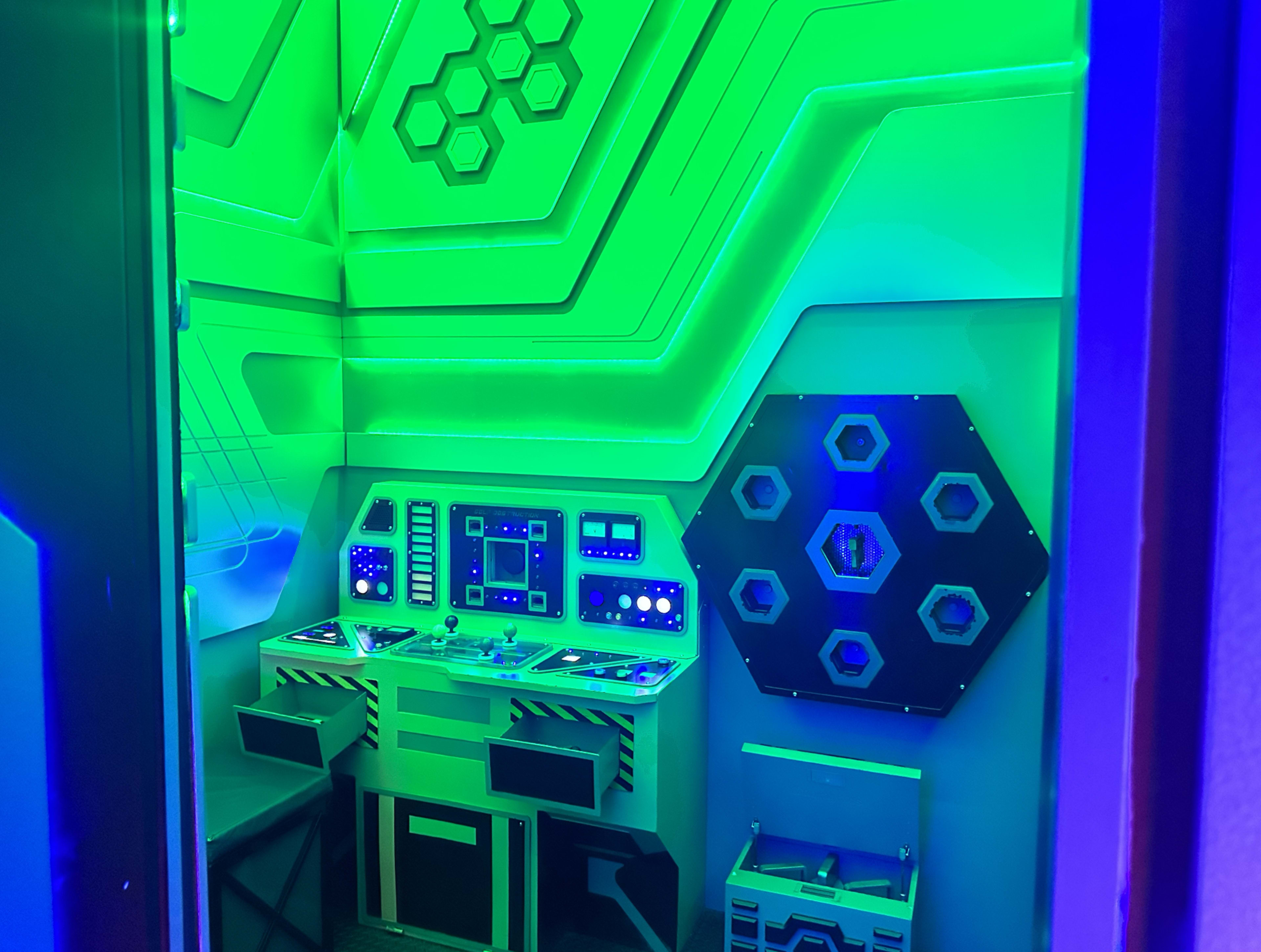 The image shows a futuristic control room with green lighting, featuring a console with buttons and screens, and hexagonal shapes on the walls.
