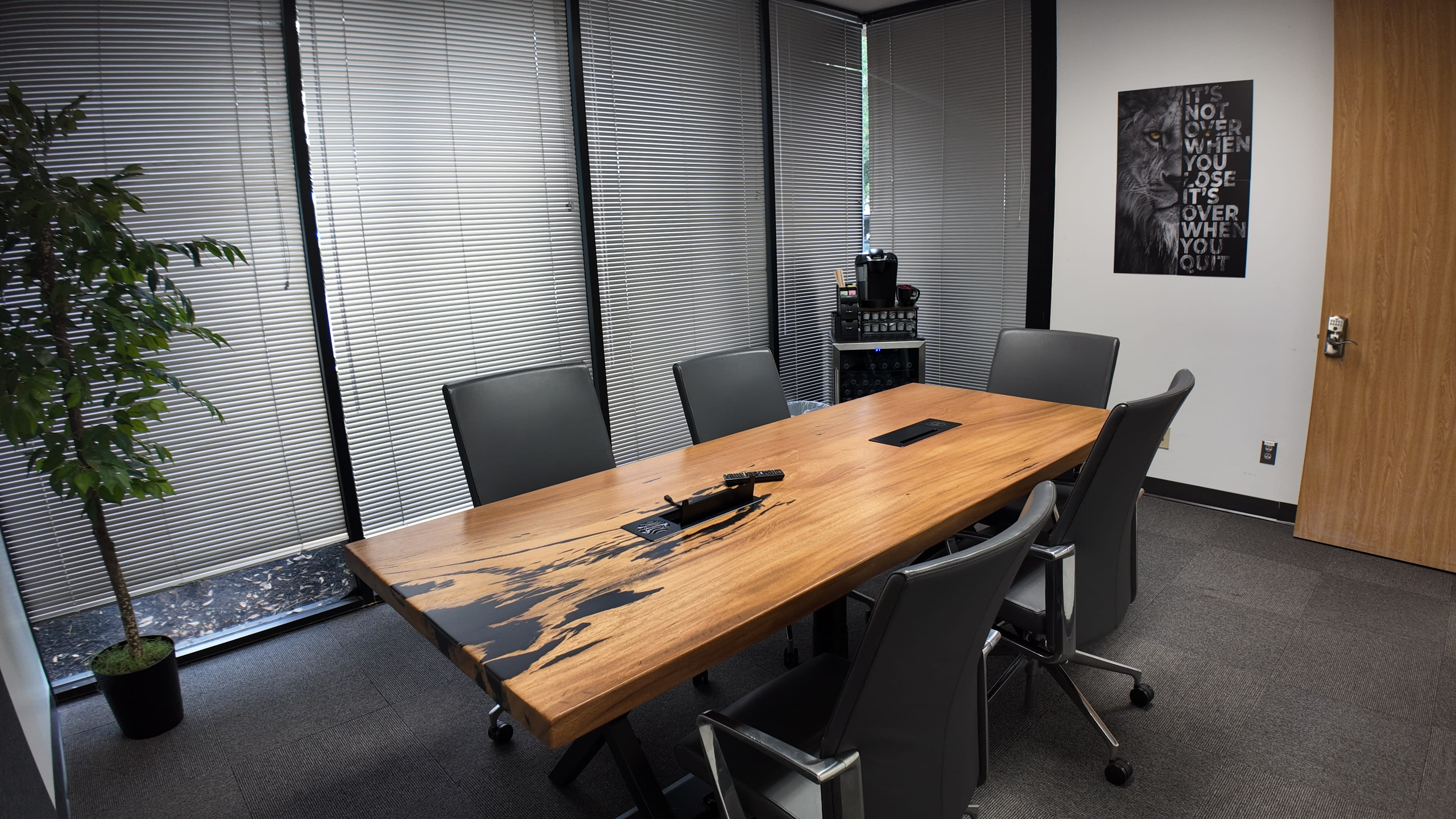 A large wooden conference table with a unique design is surrounded by six gray office chairs in a well-lit meeting room with window blinds.