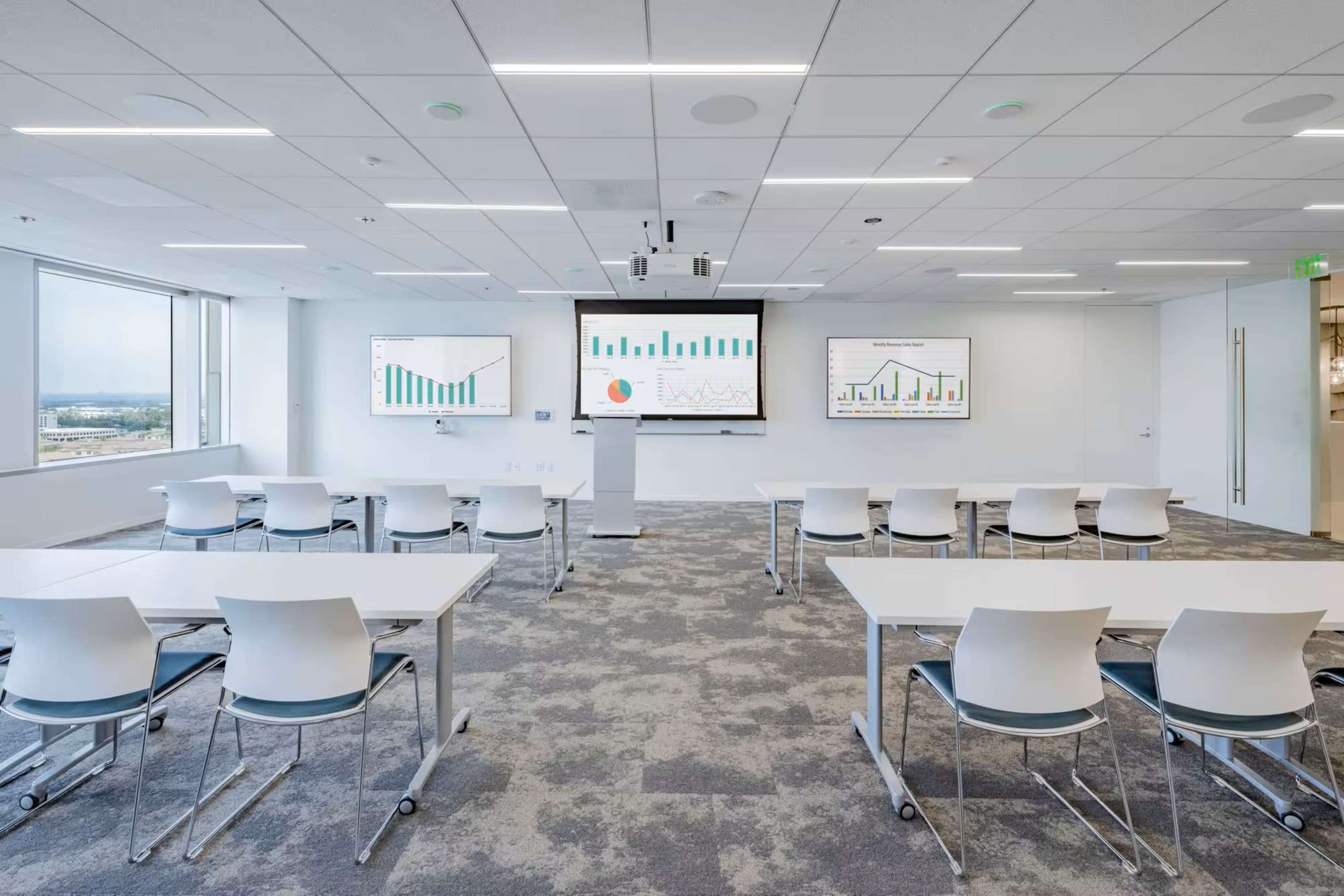 A modern conference room features multiple screens displaying graphs, arranged seating with white tables and chairs, and large windows providing natural light.