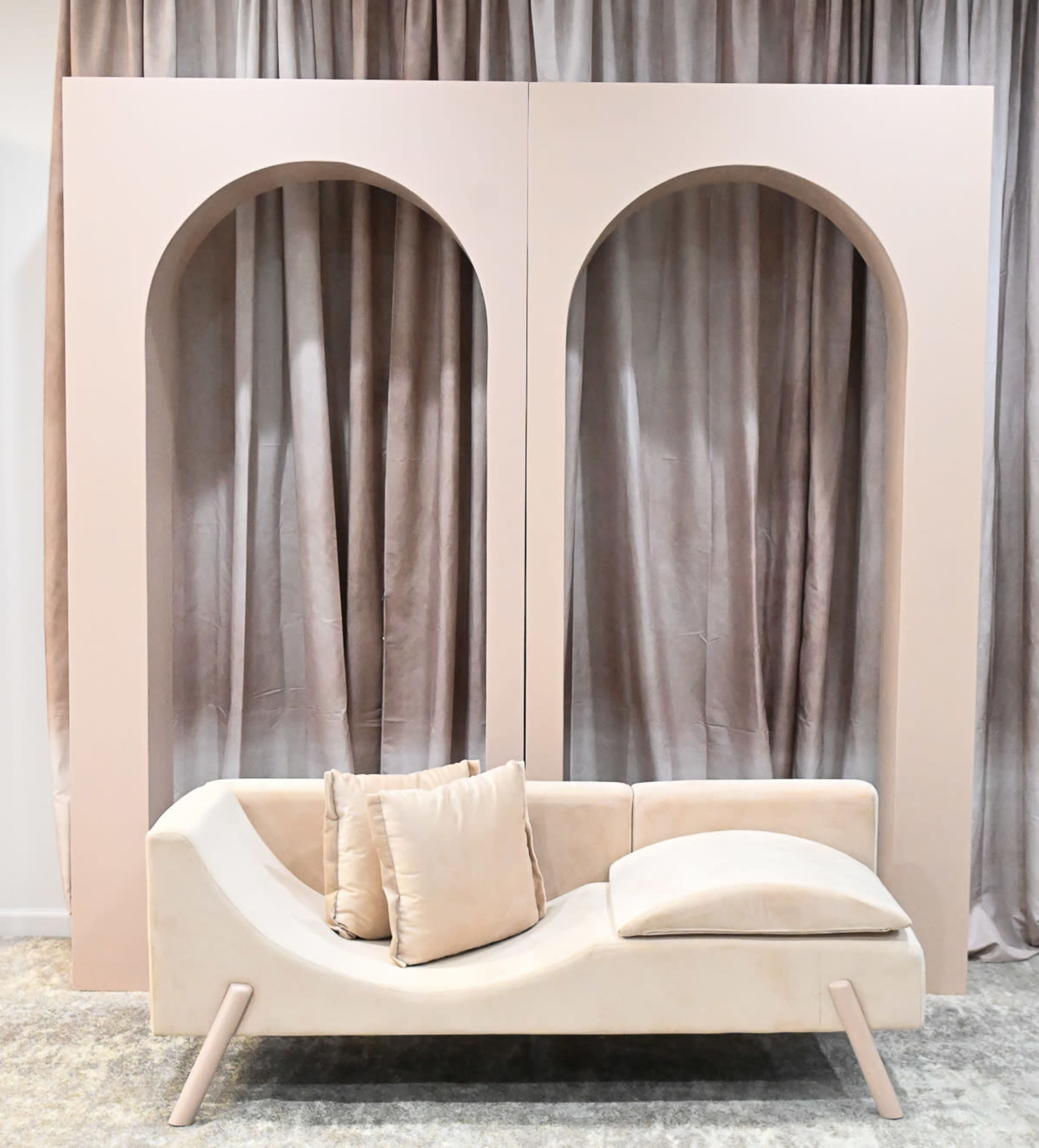 A sleek, curved sofa with a light fabric is positioned in front of a backdrop featuring two arch-shaped cutouts and soft, pleated curtains.