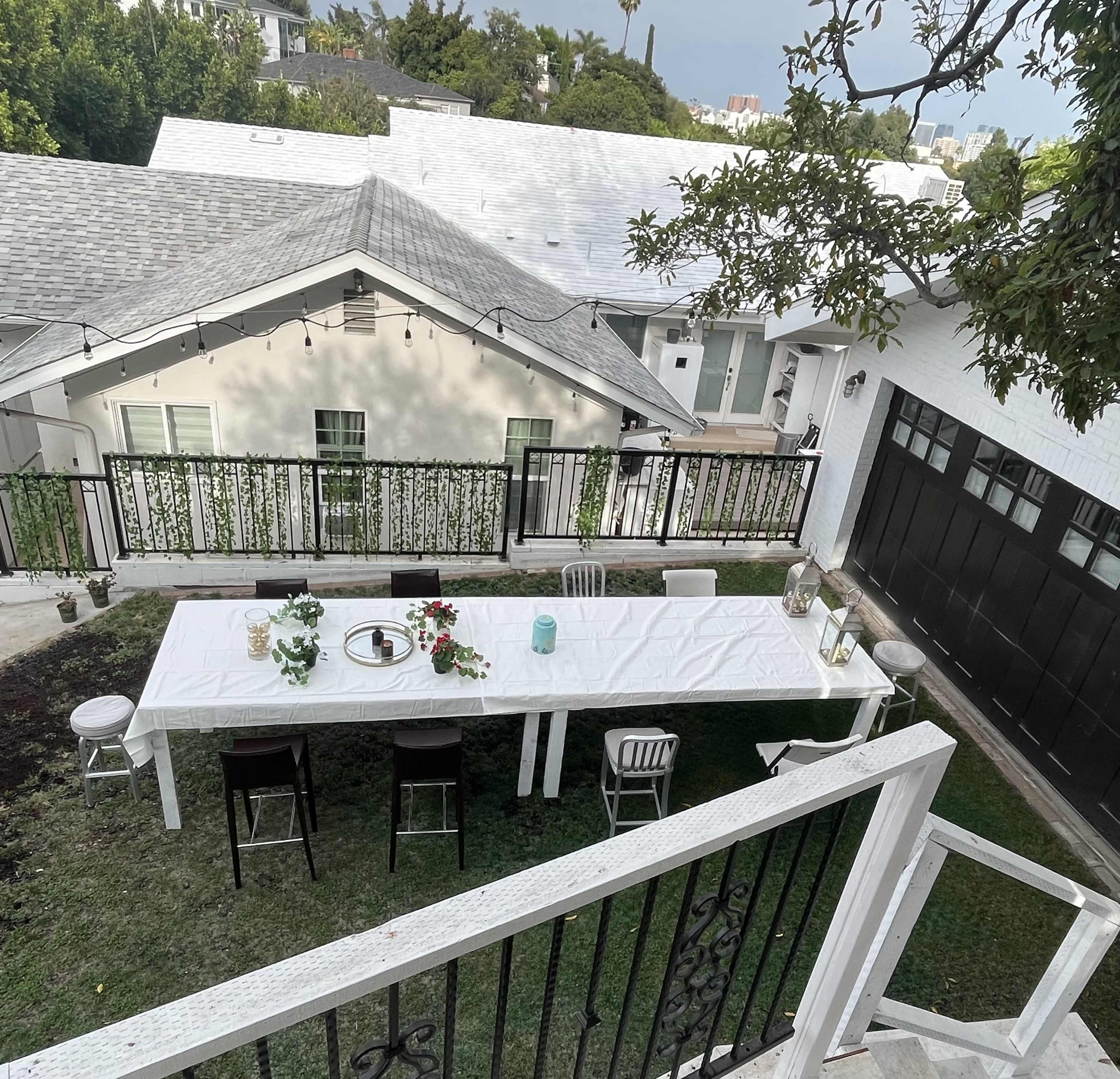 A long white table is set on grass in front of a house, surrounded by chairs and decorated with plants and tableware.