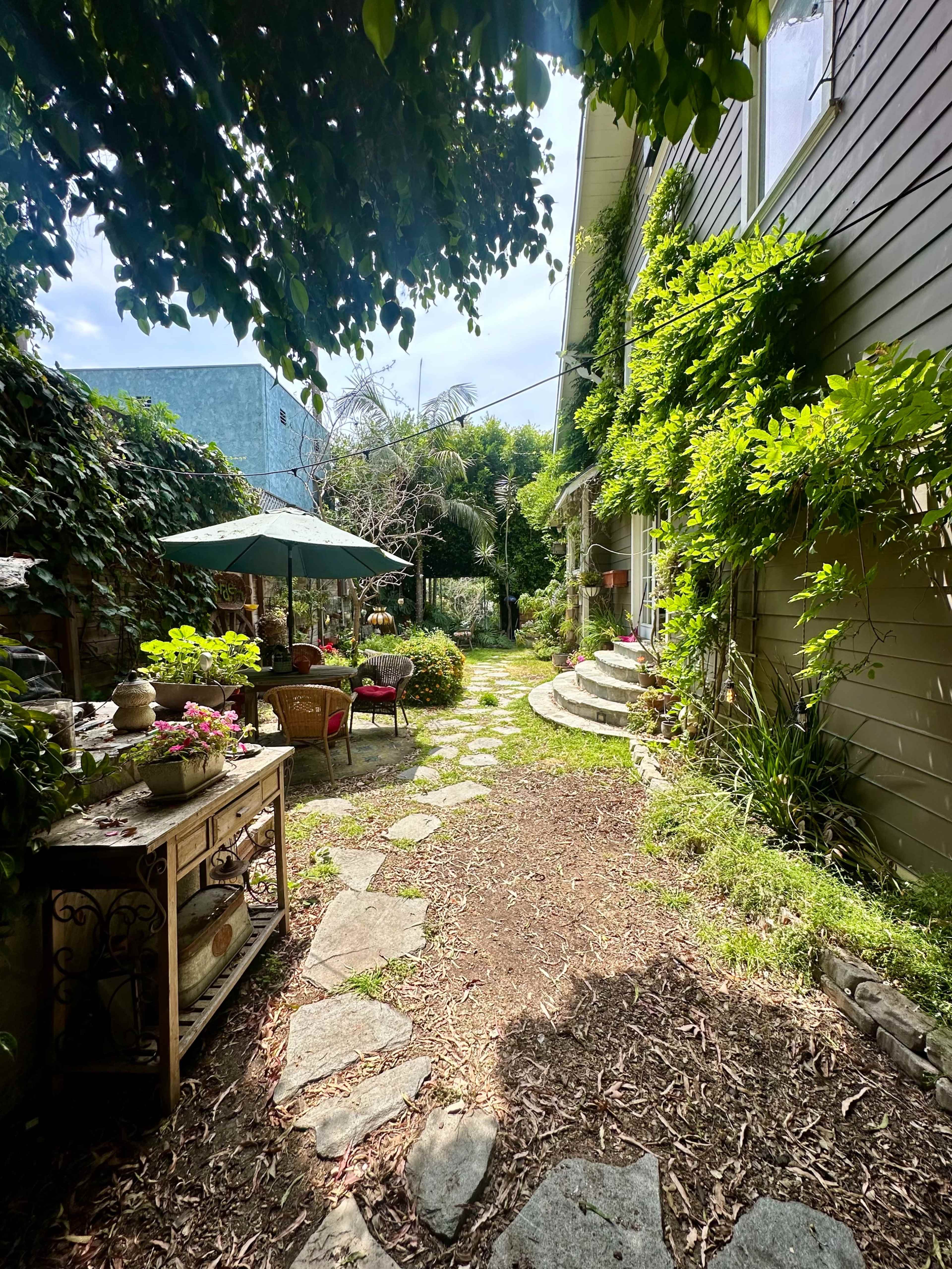 A stone pathway leads through a lush garden, flanked by greenery and decorative furniture, toward a house.