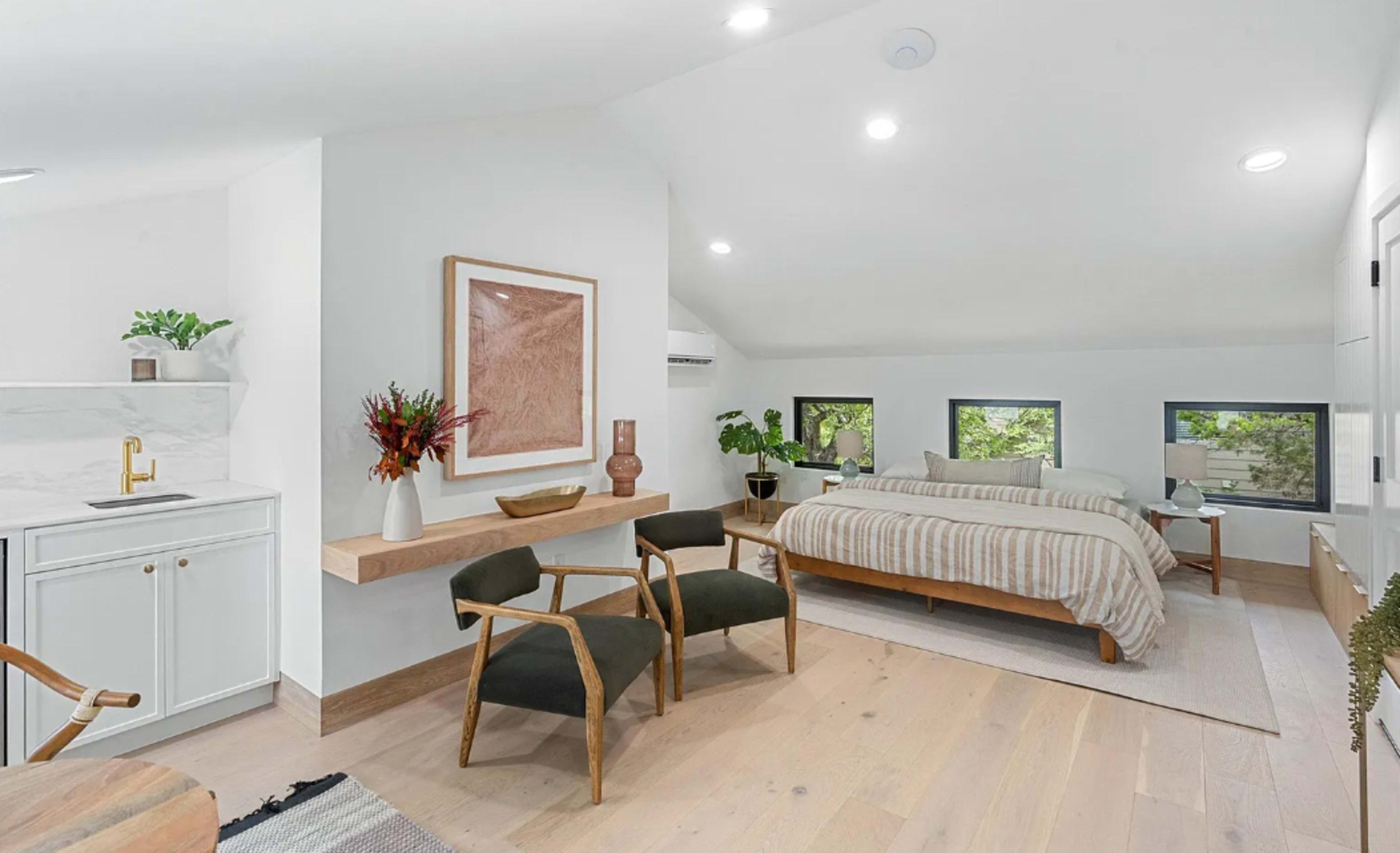The image depicts a modern bedroom with a neatly made bed, a small kitchenette, and two chairs, all set against a backdrop of large windows allowing natural light to enter.