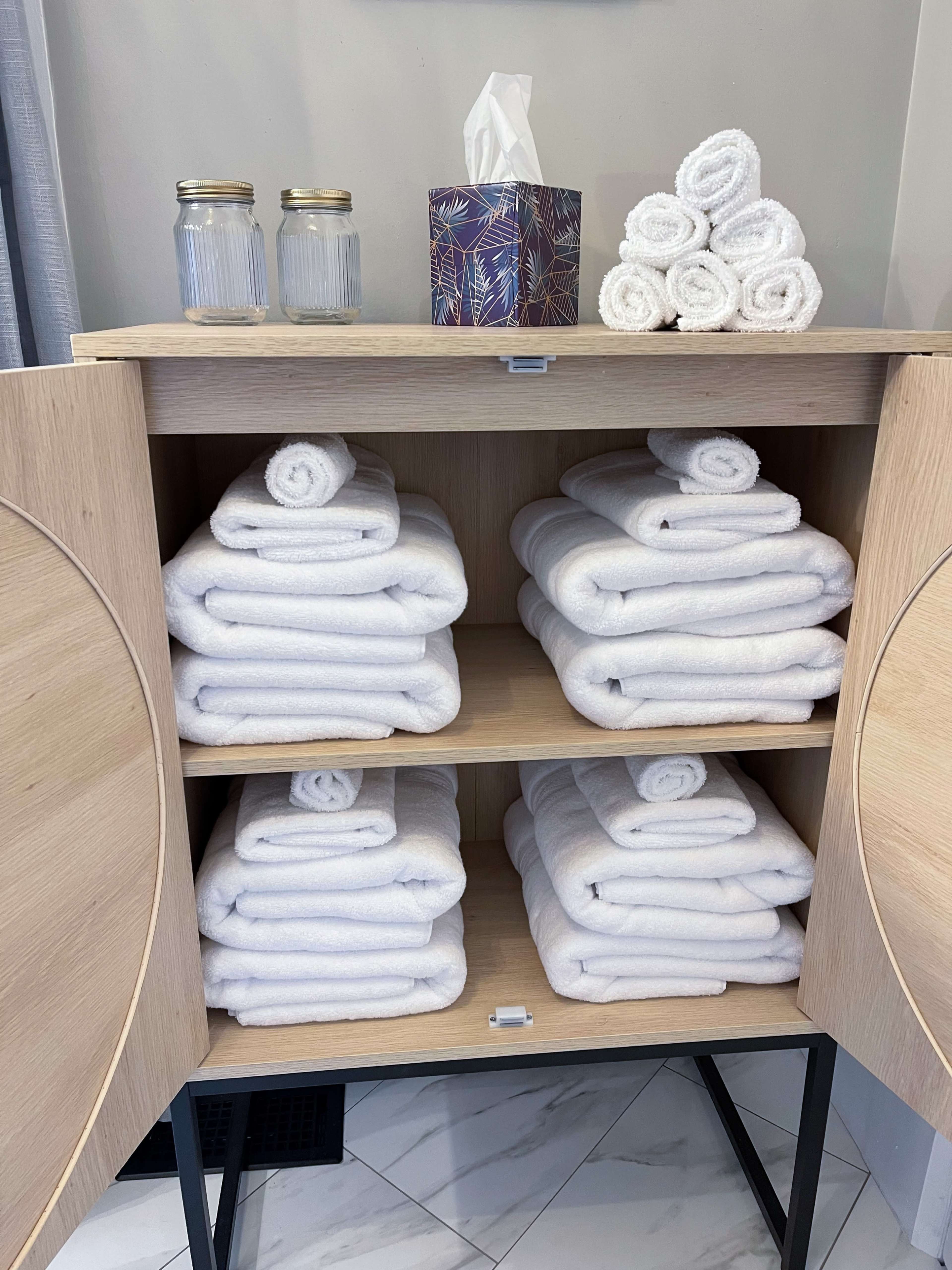 A wooden cabinet with neatly arranged white towels on its shelves, along with a box of tissues and two glass jars on top.