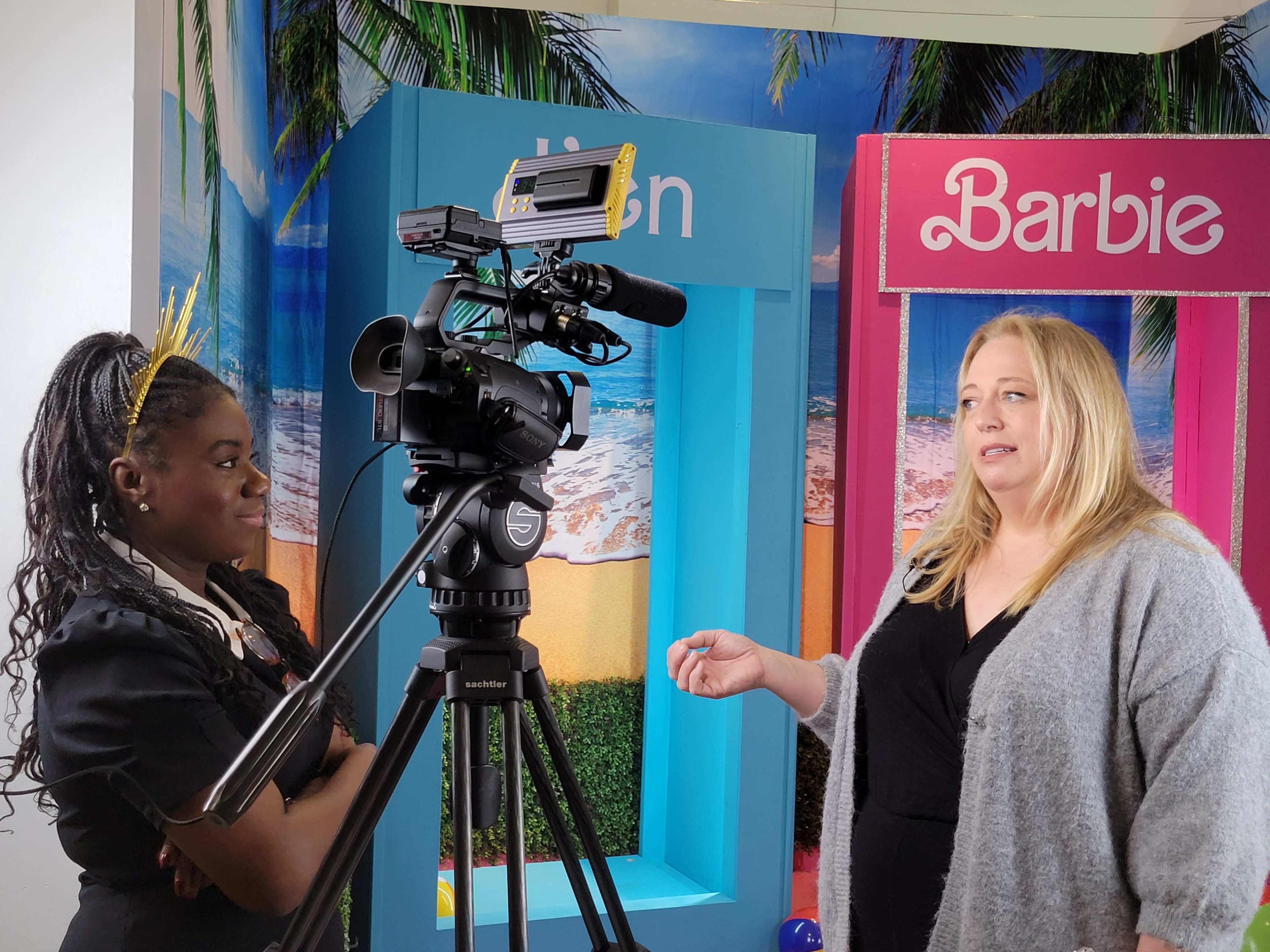 A woman is being interviewed in front of colorful backdrops featuring the names "Elon" and "Barbie," while another person operates a camera.
