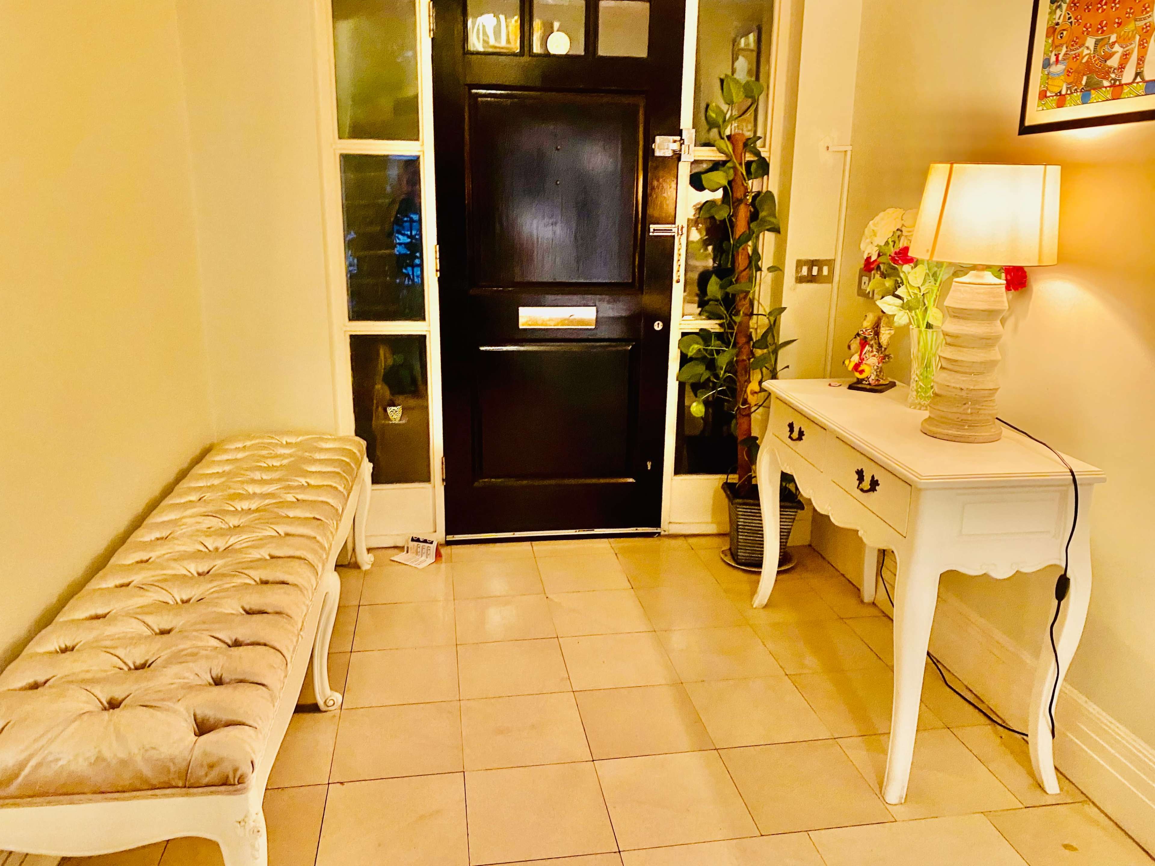 A hallway with a black door, a tufted bench, a small white console table with a decorative lamp, and a plant in the corner.