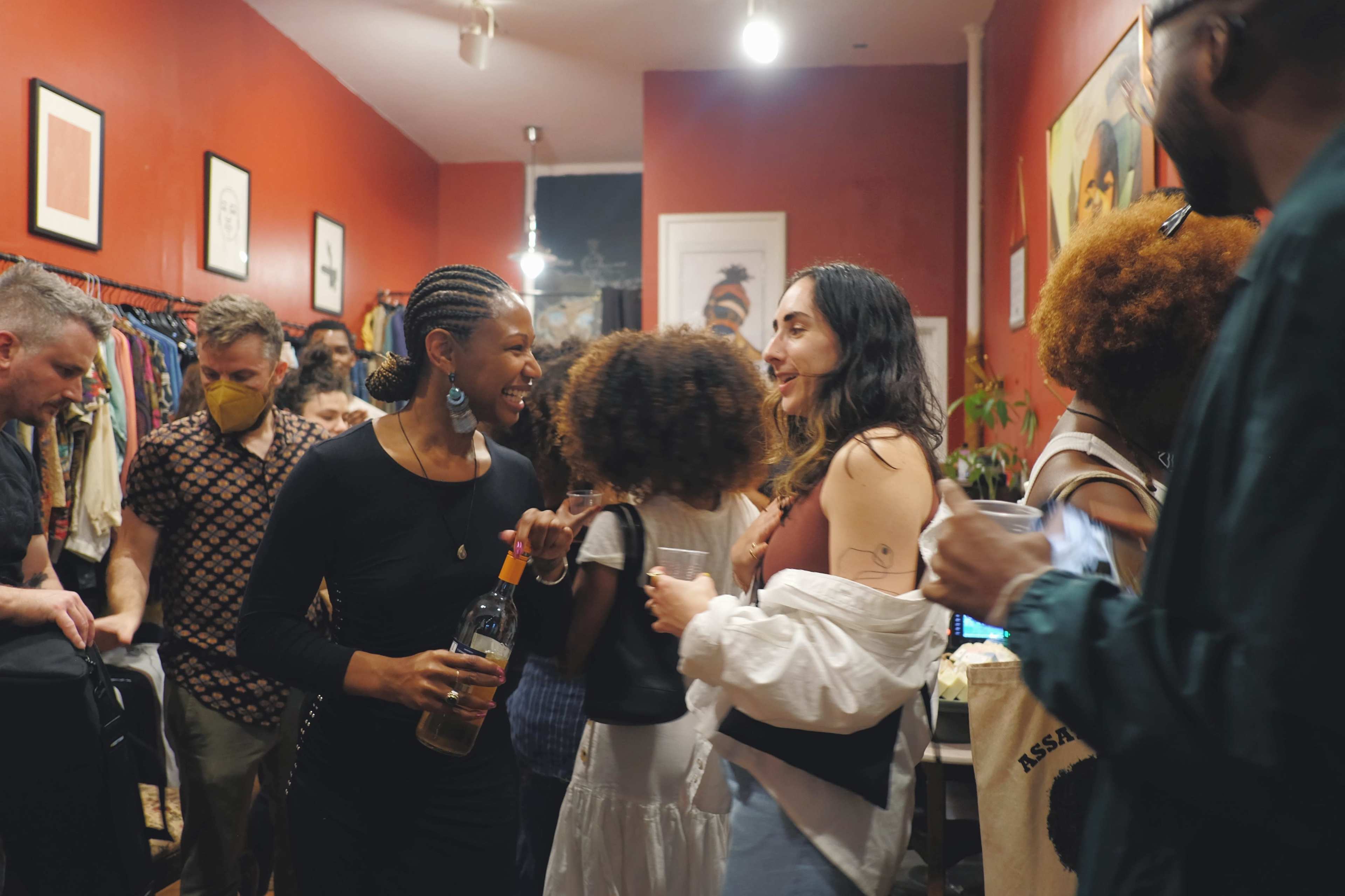 A diverse group of people socializes in a vintage clothing store, surrounded by colorful racks of clothing and art on the walls.