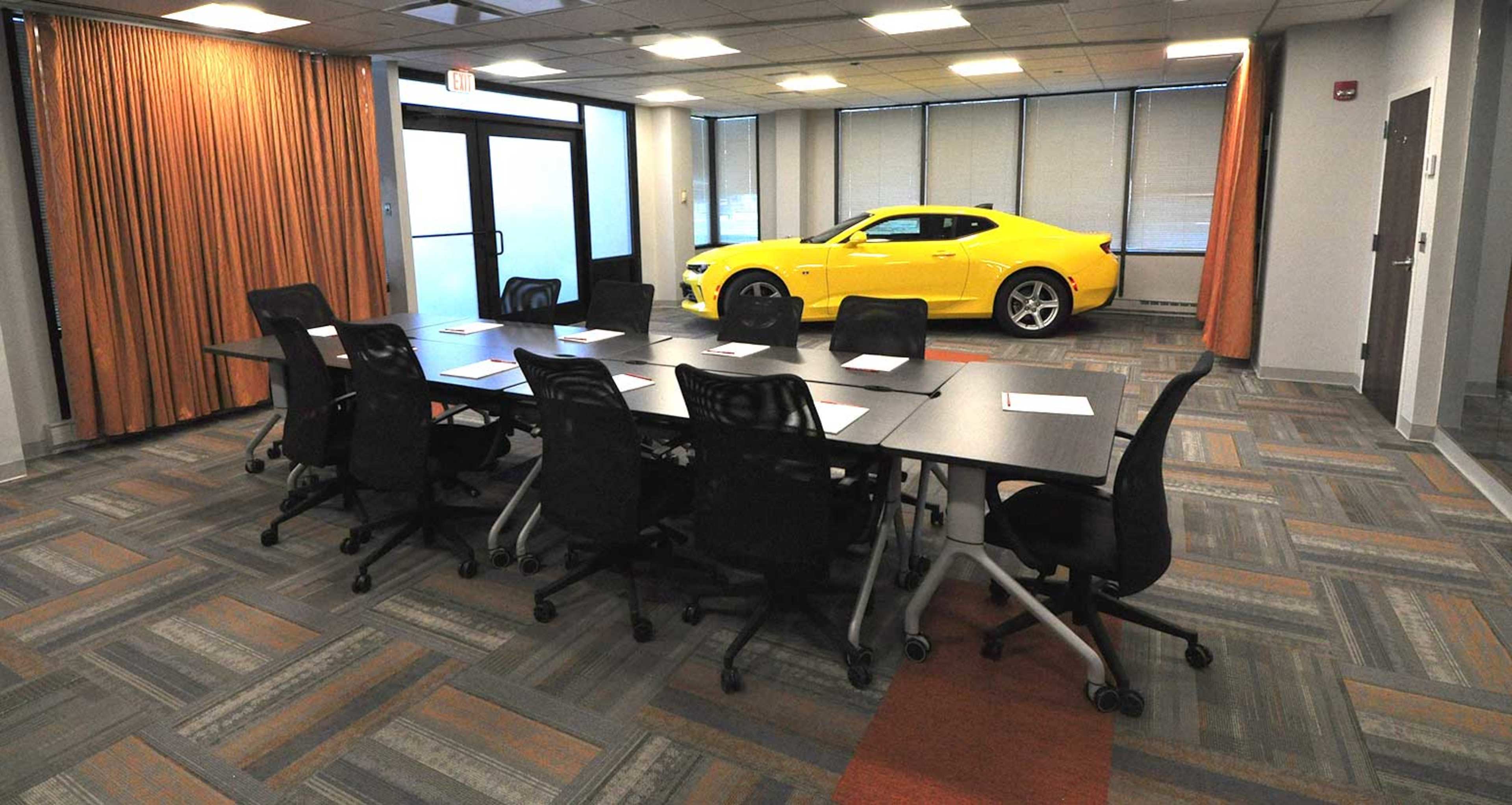 A conference room features a long table surrounded by chairs, with a yellow sports car parked beside large windows.