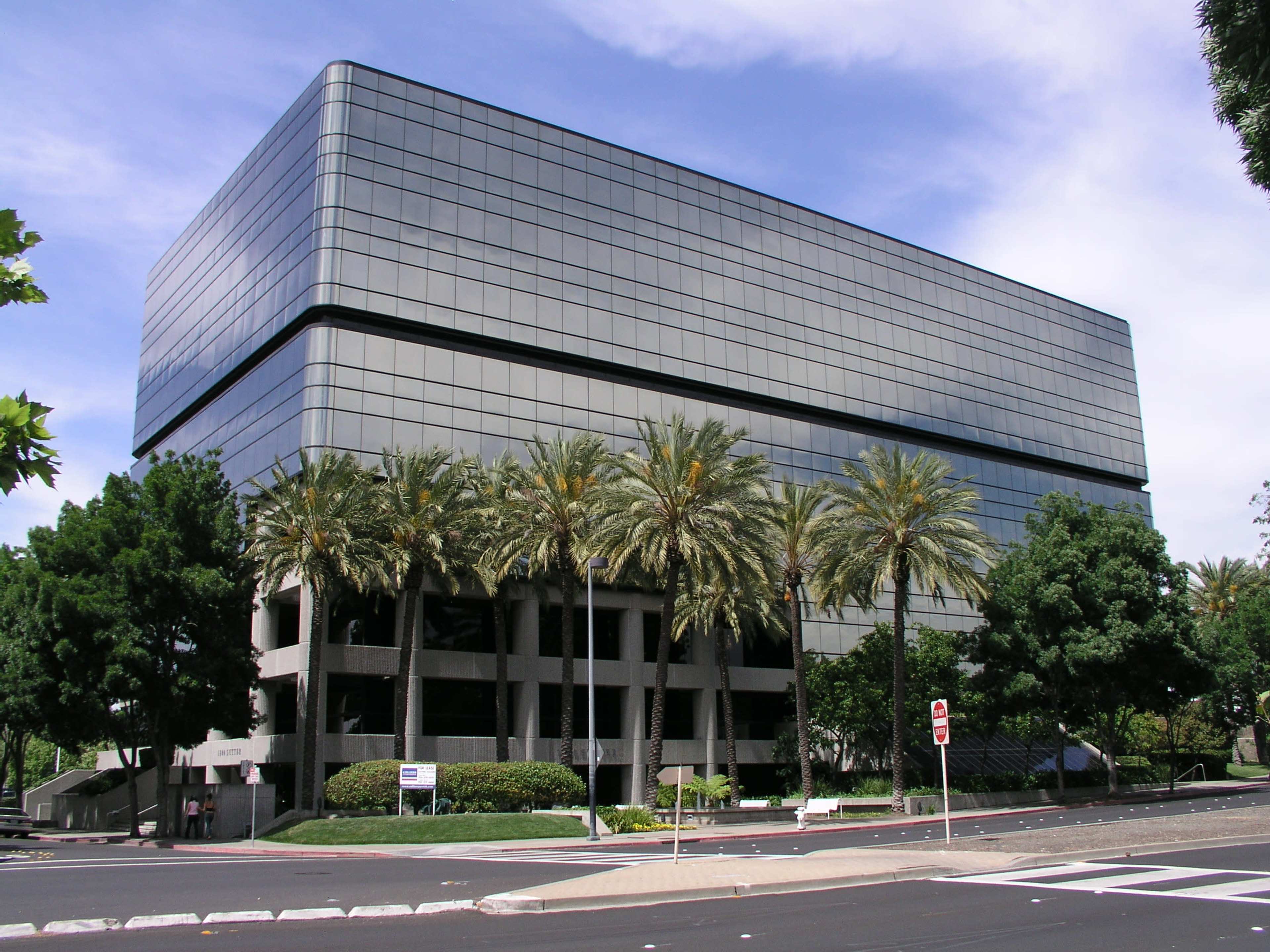 A modern, glass-fronted building with a palm-lined entrance is situated at the intersection of two streets.