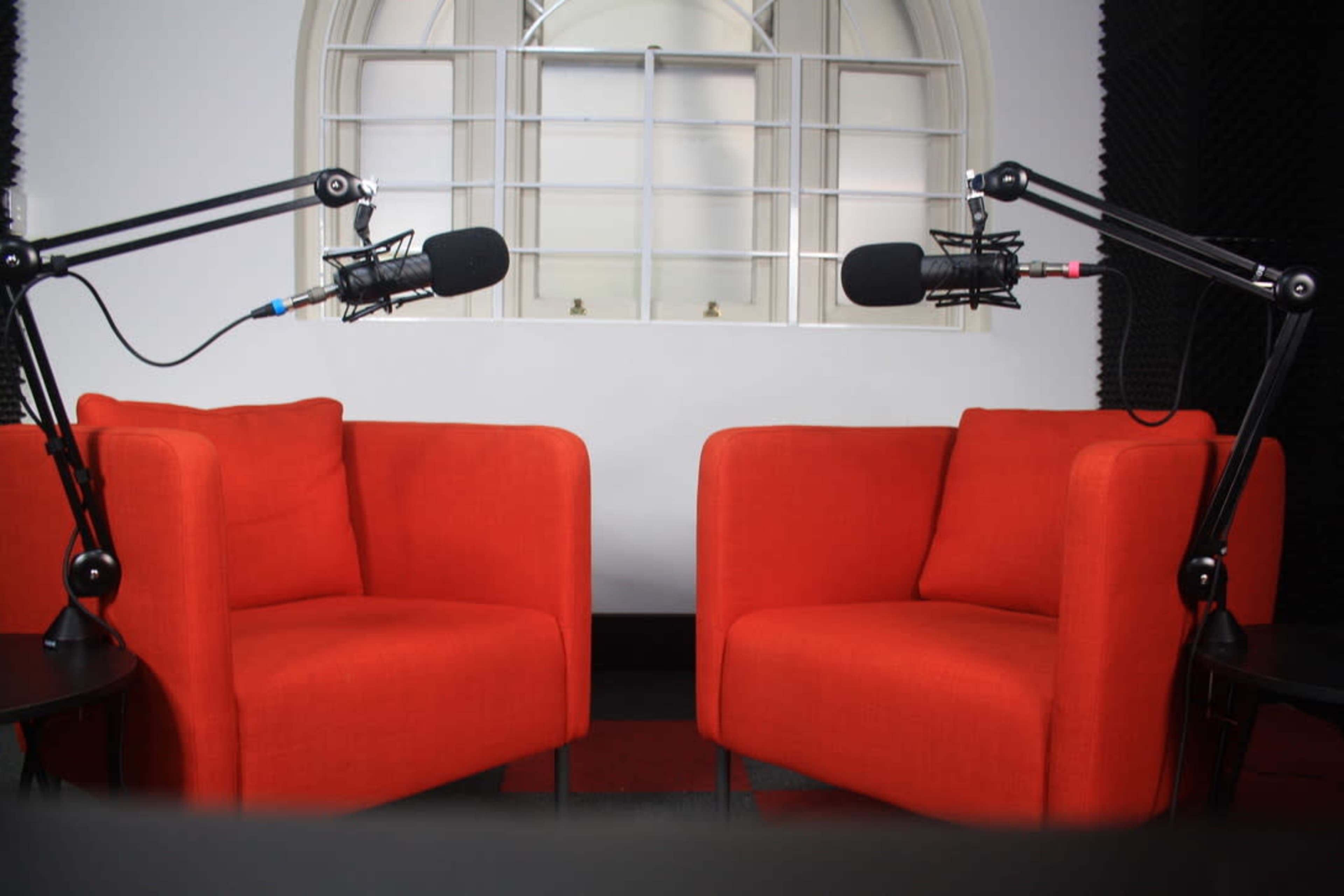 Two red armchairs positioned facing each other, each equipped with a microphone on an adjustable stand, set against a wall with a large window.