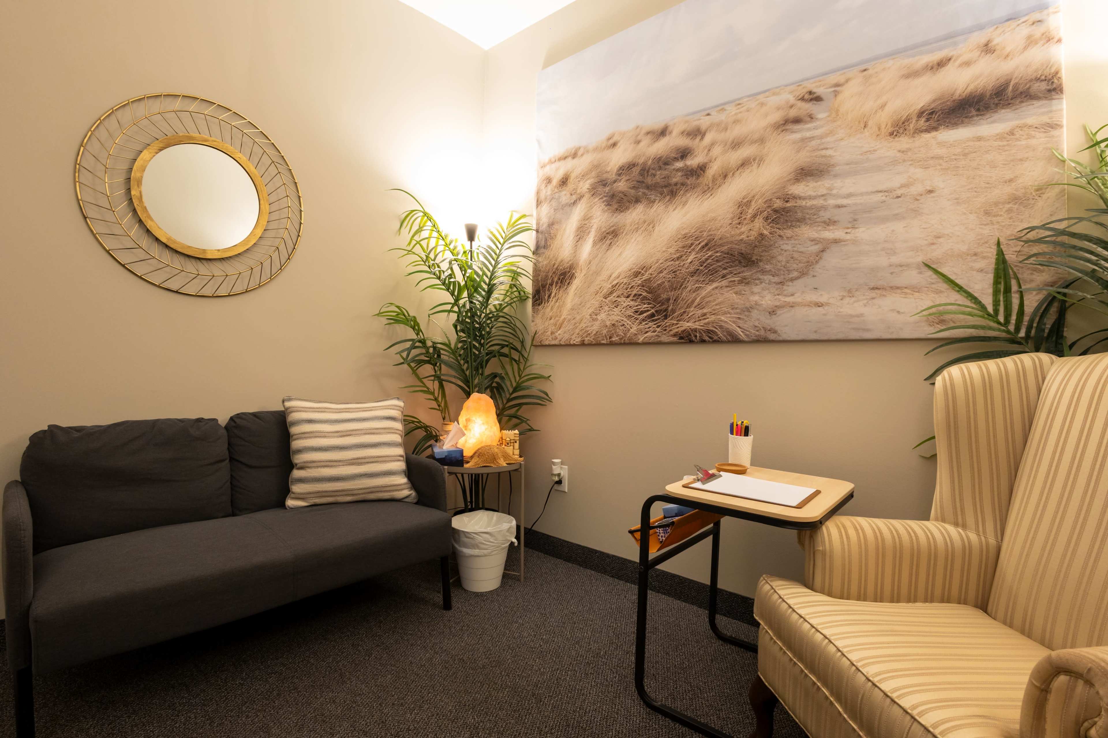 The image shows a small, cozy therapy room with a gray couch, an armchair, a side table, a Himalayan salt lamp, and a large nature-themed wall mural.