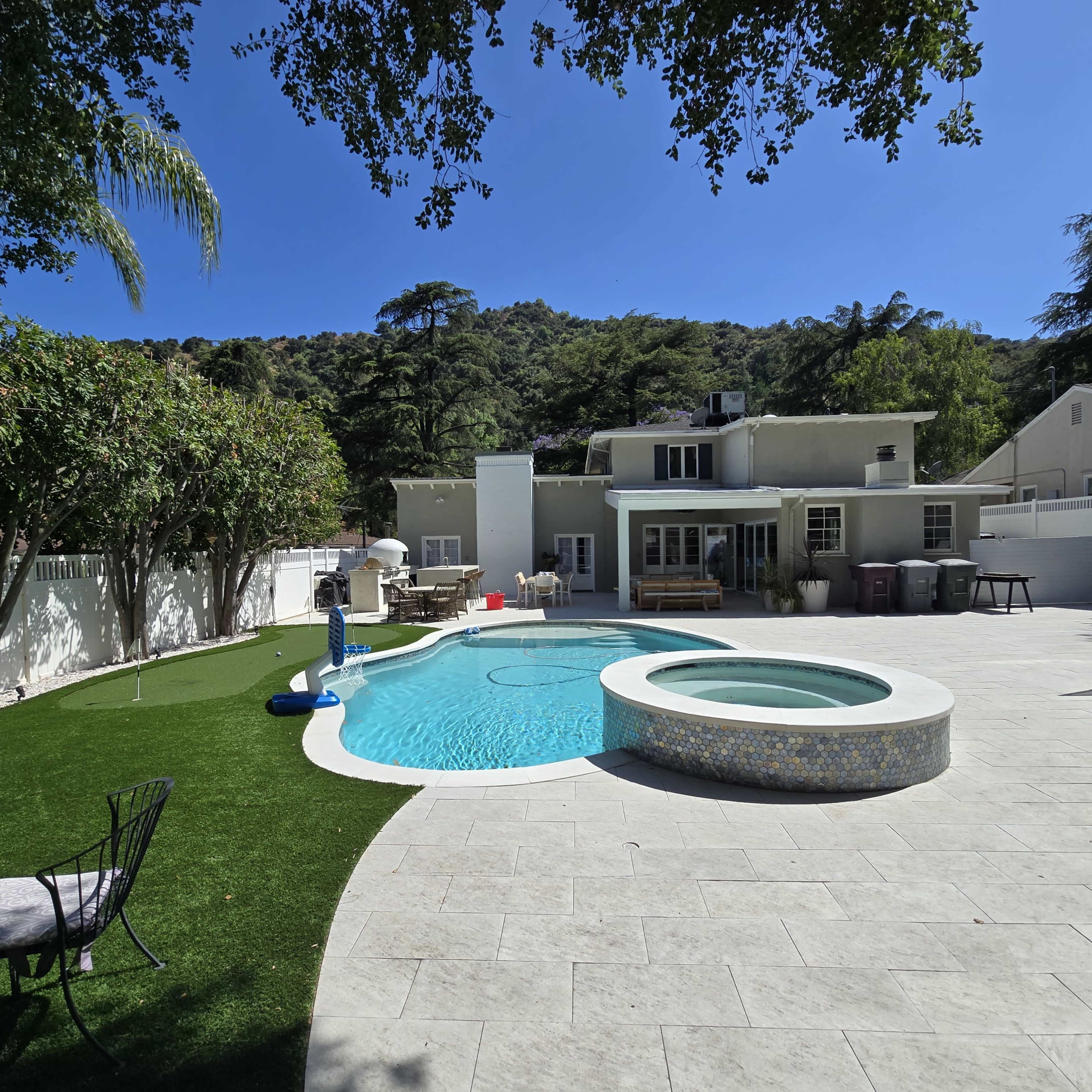 The image shows a modern backyard featuring a swimming pool with an attached spa, surrounded by a patio area, artificial grass, and a hillside in the background.