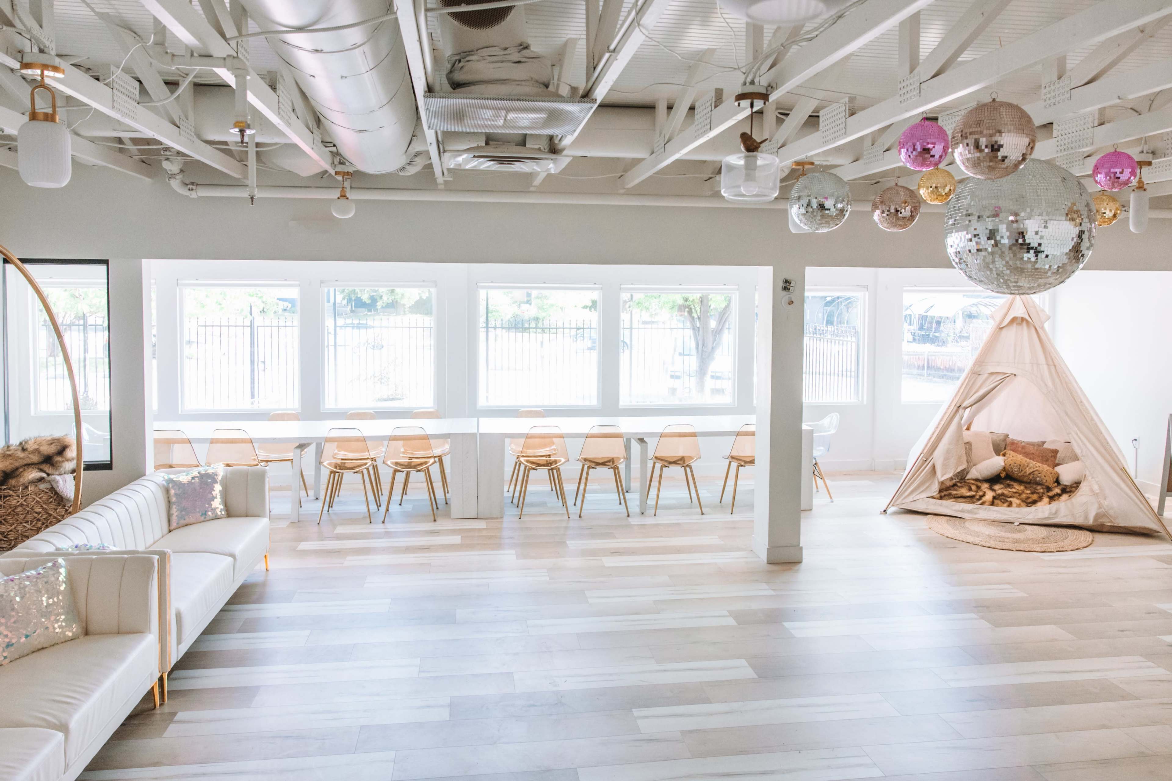A modern, airy interior space with white walls, light wooden flooring, a long table with chairs, a seating area, and a decorative teepee.