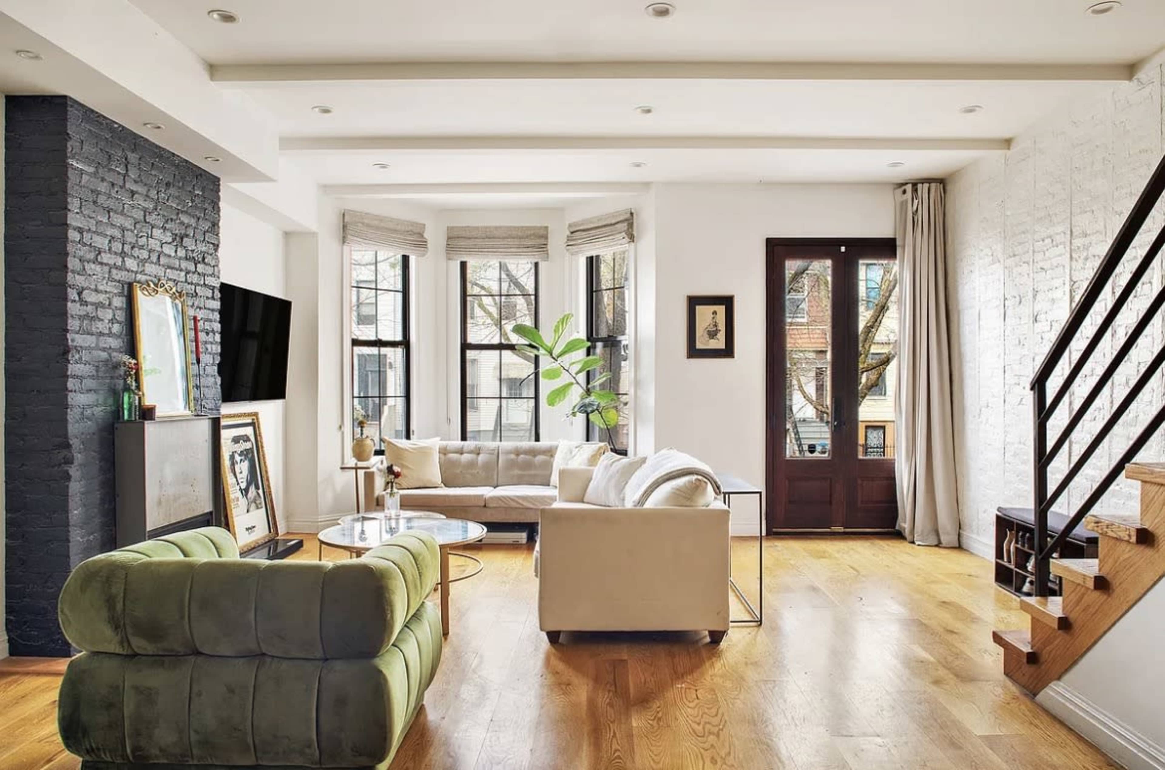 The image shows a spacious living room with a mix of modern furniture, large windows, and a staircase leading to an upper level.