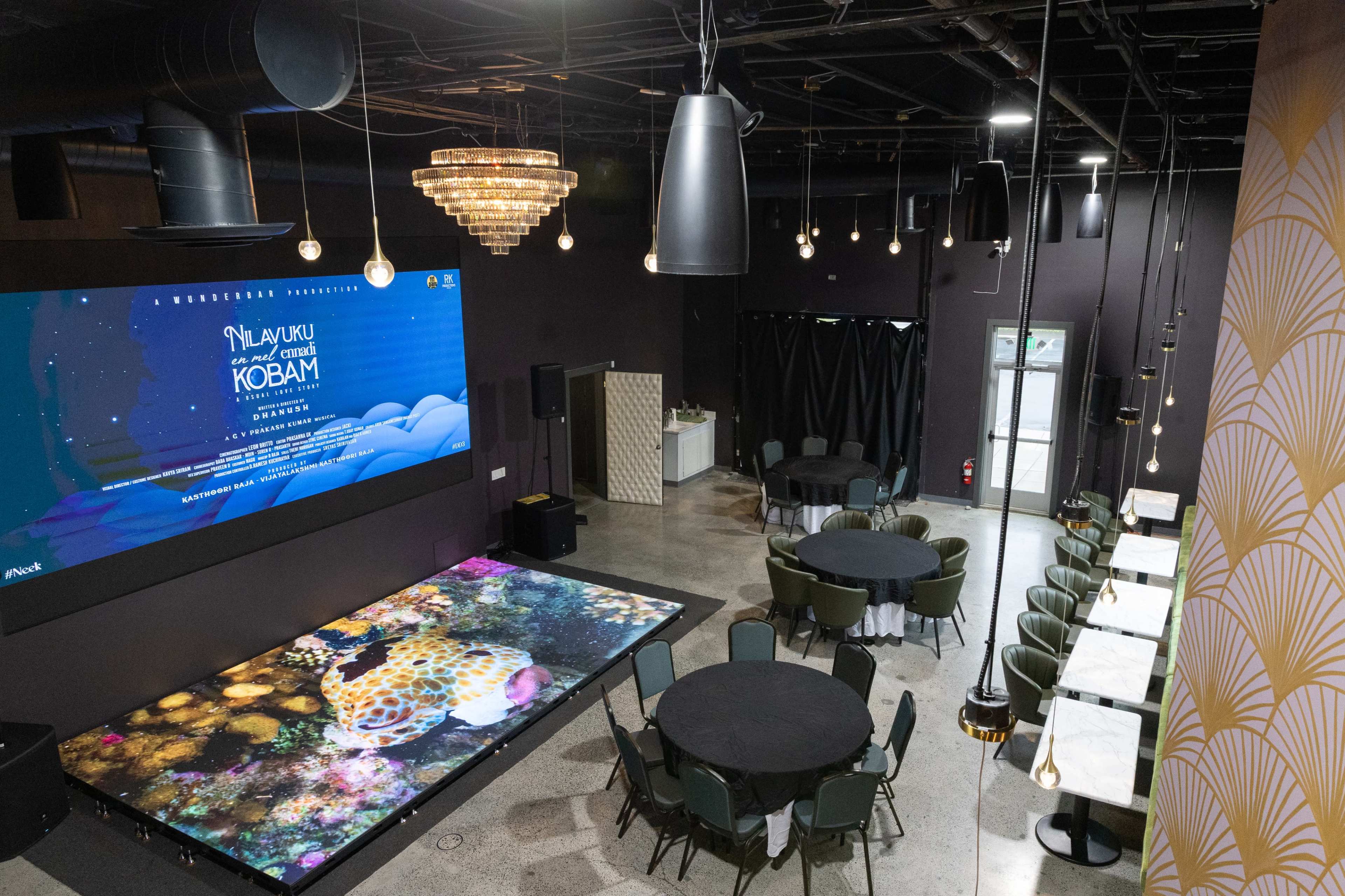 The image depicts a modern event space featuring a large digital screen and seating arrangements with round tables and decorative lighting.