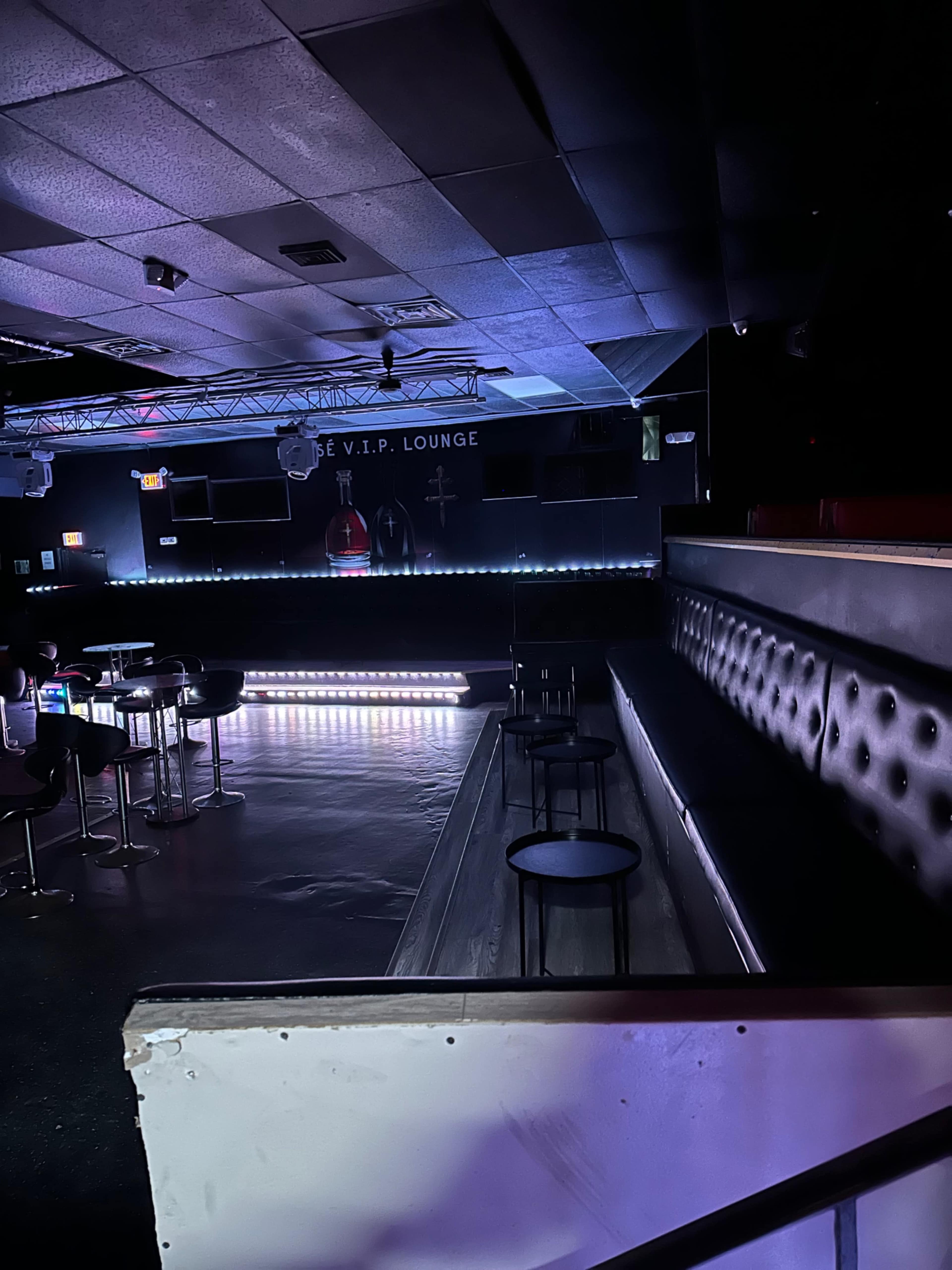 The image shows the interior of a dimly lit lounge with black seating, high tables, and a small stage area in the background.