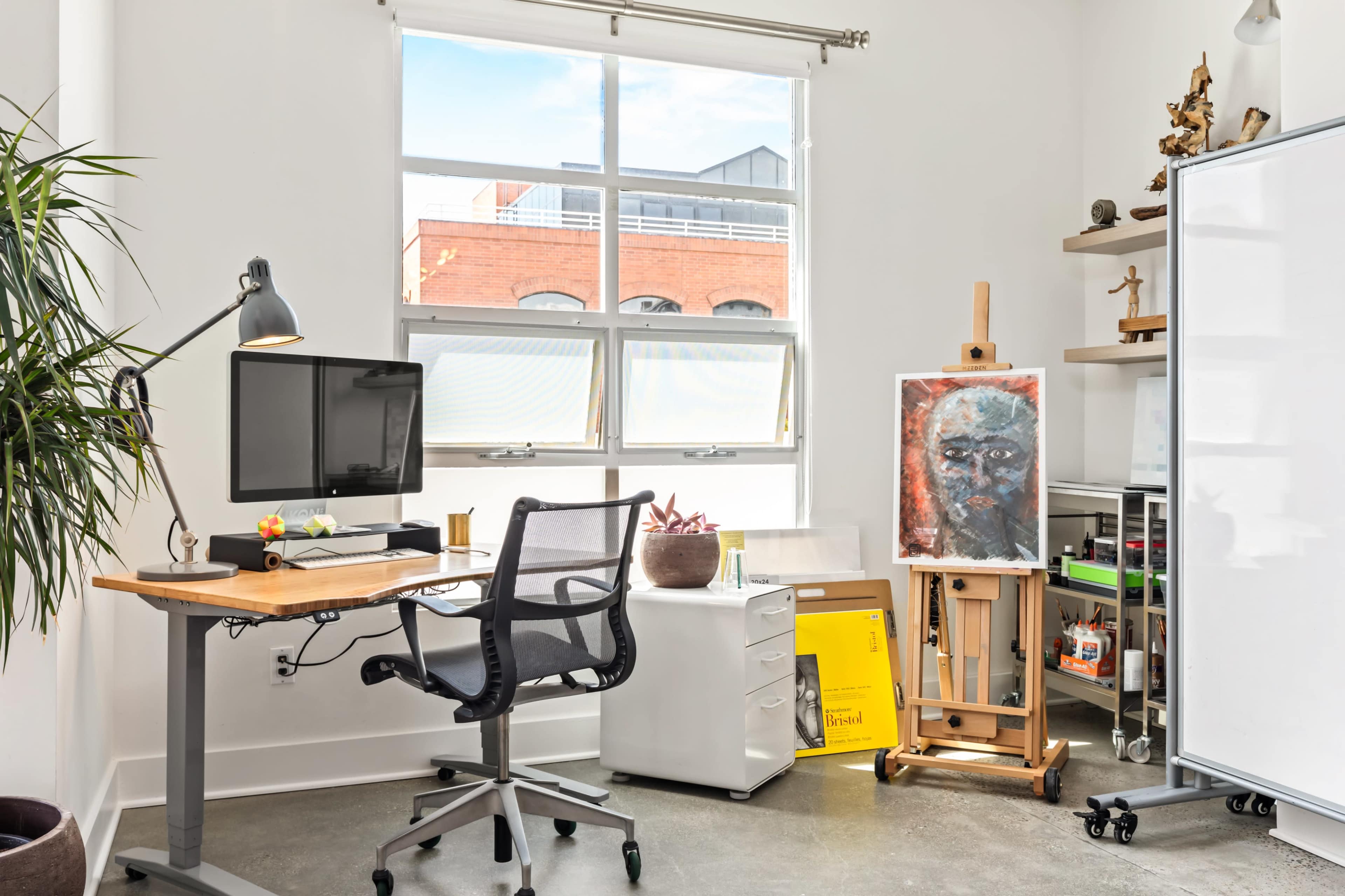 The image shows a modern office space with a desk, computer, and an easel displaying an artwork, along with a potted plant and storage units.