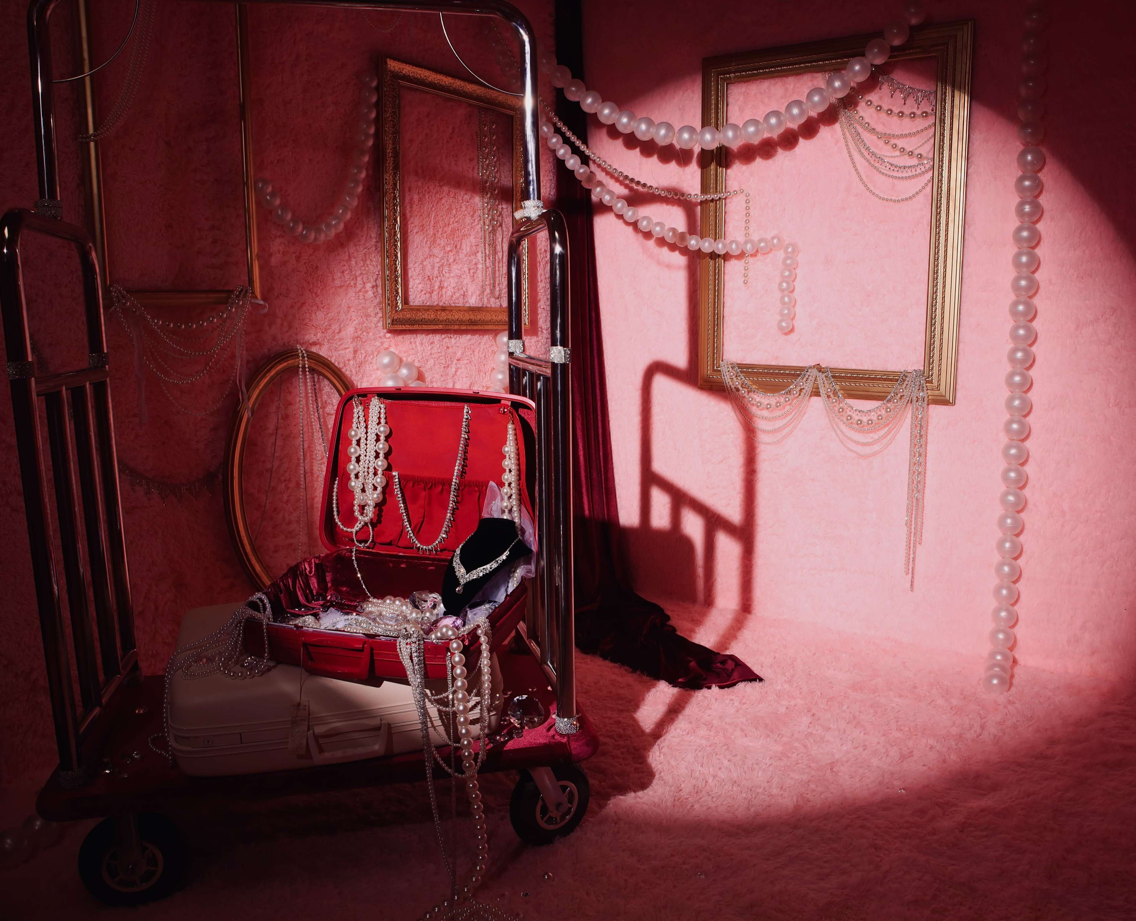 A red luggage cart displays a red suitcase filled with various strands of pearls and jewelry against a pink background adorned with framed mirrors and draped pearls.