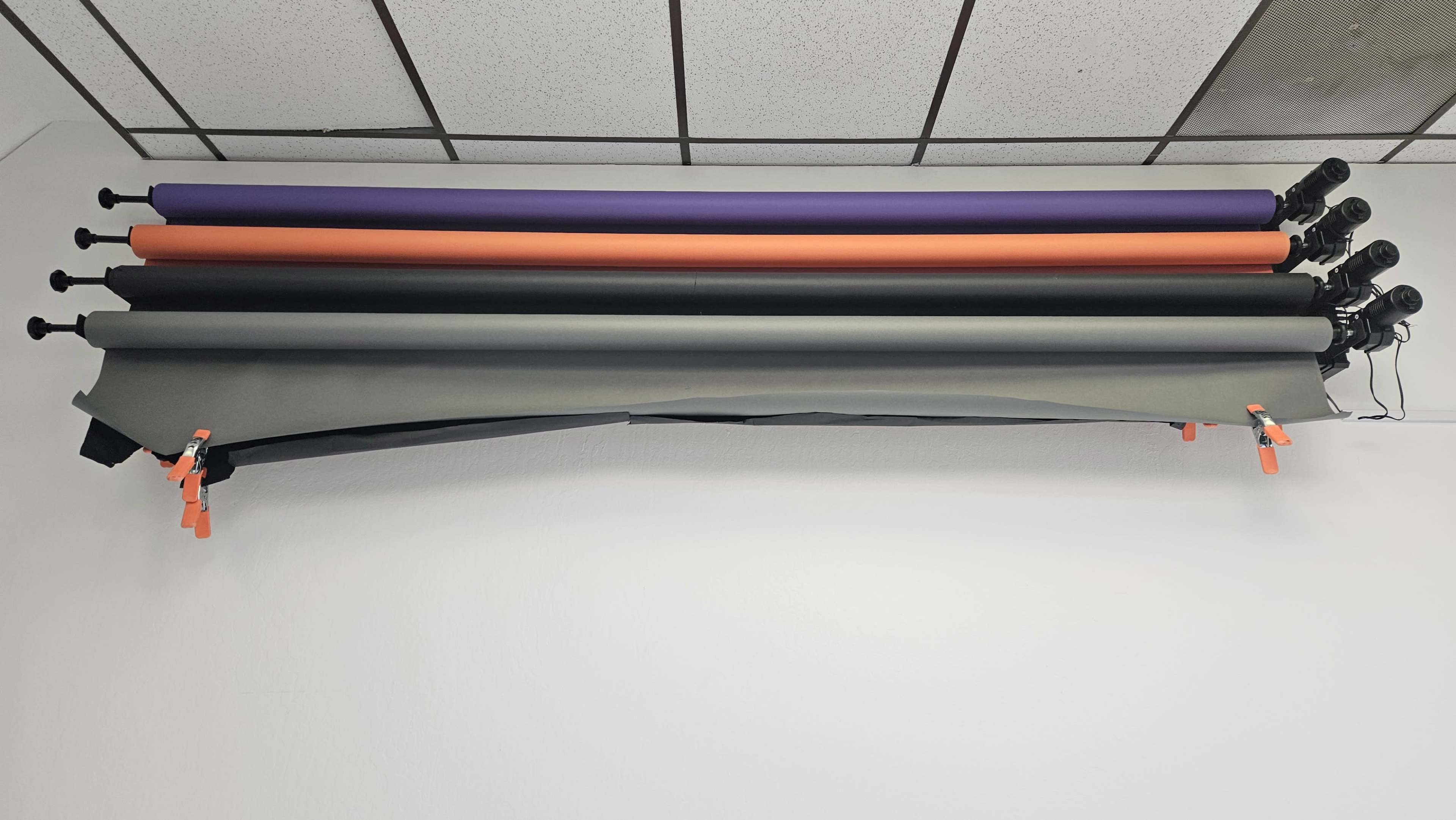 Several rolls of background paper, including gray, orange, purple, and black, mounted on a horizontal bar and secured with clamps on a wall.
