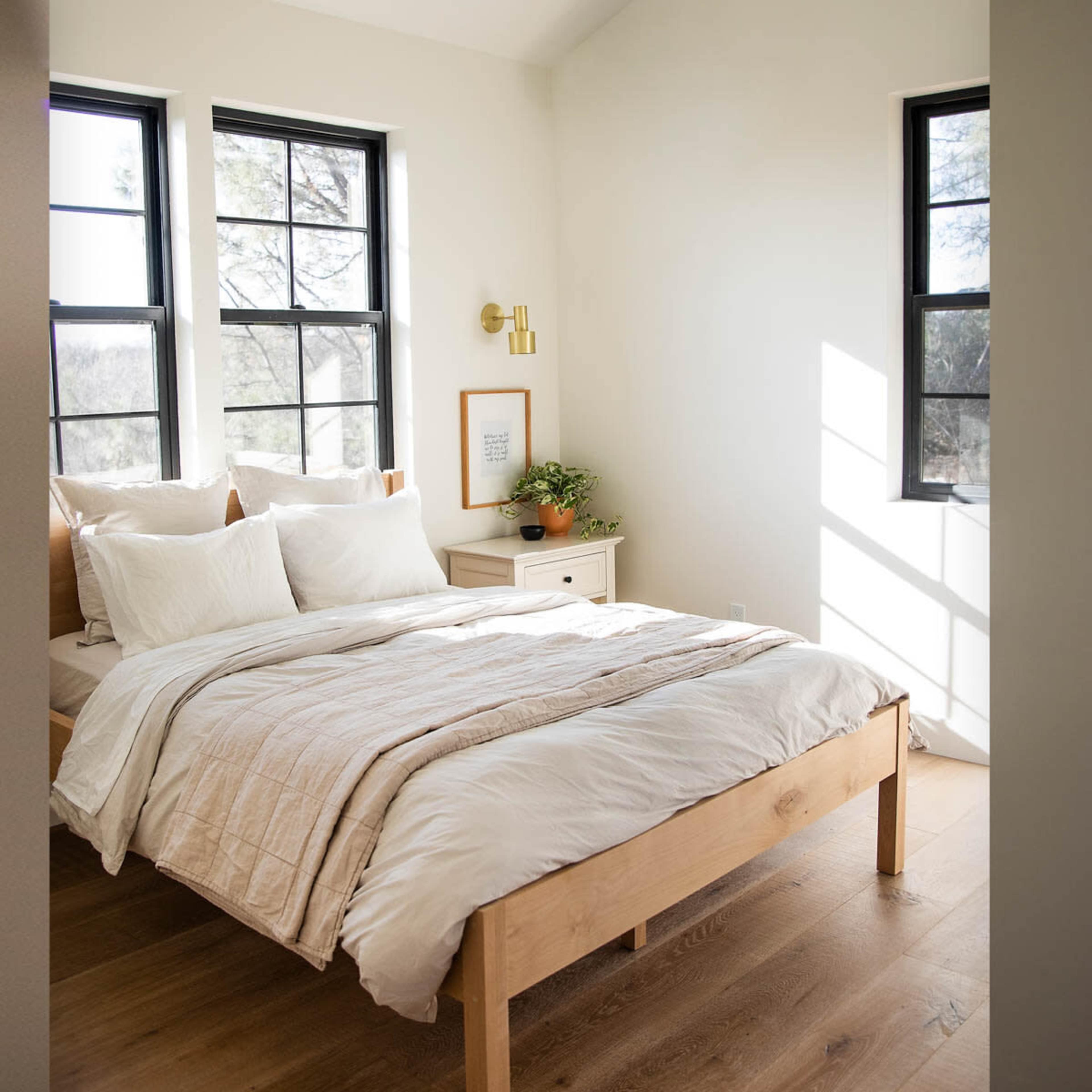 A well-lit bedroom features a wooden bed with a light-colored comforter and two windows allowing natural light, along with a nightstand and a potted plant.