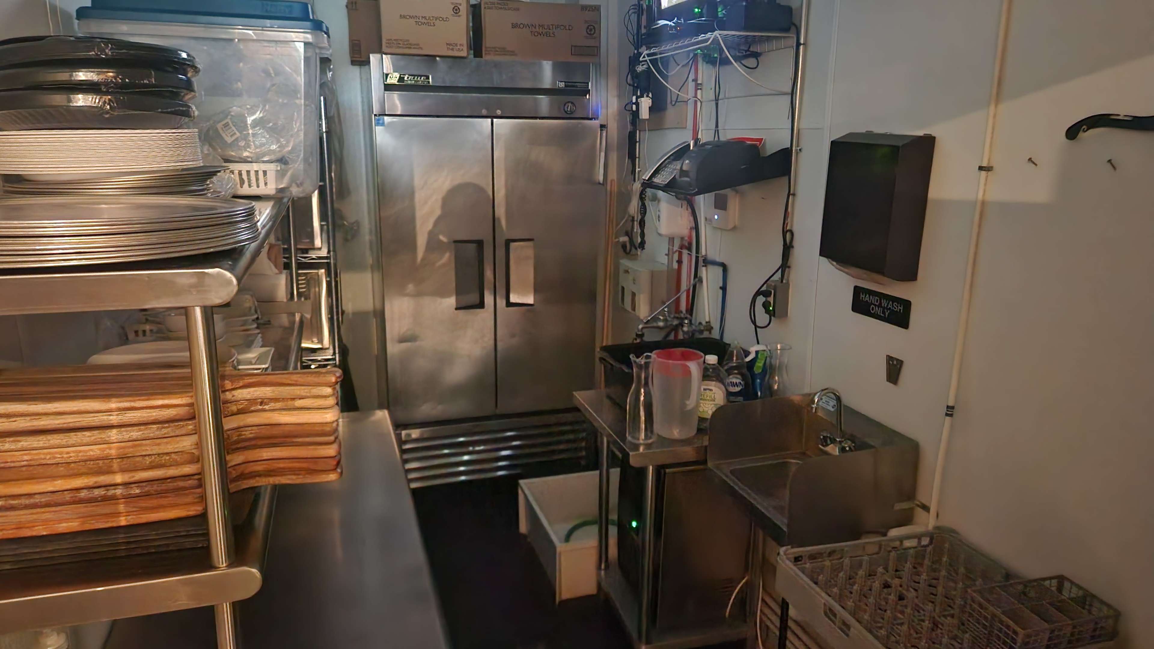 A commercial kitchen with stainless steel appliances, shelves stacked with plates, and a large refrigerator in the background.