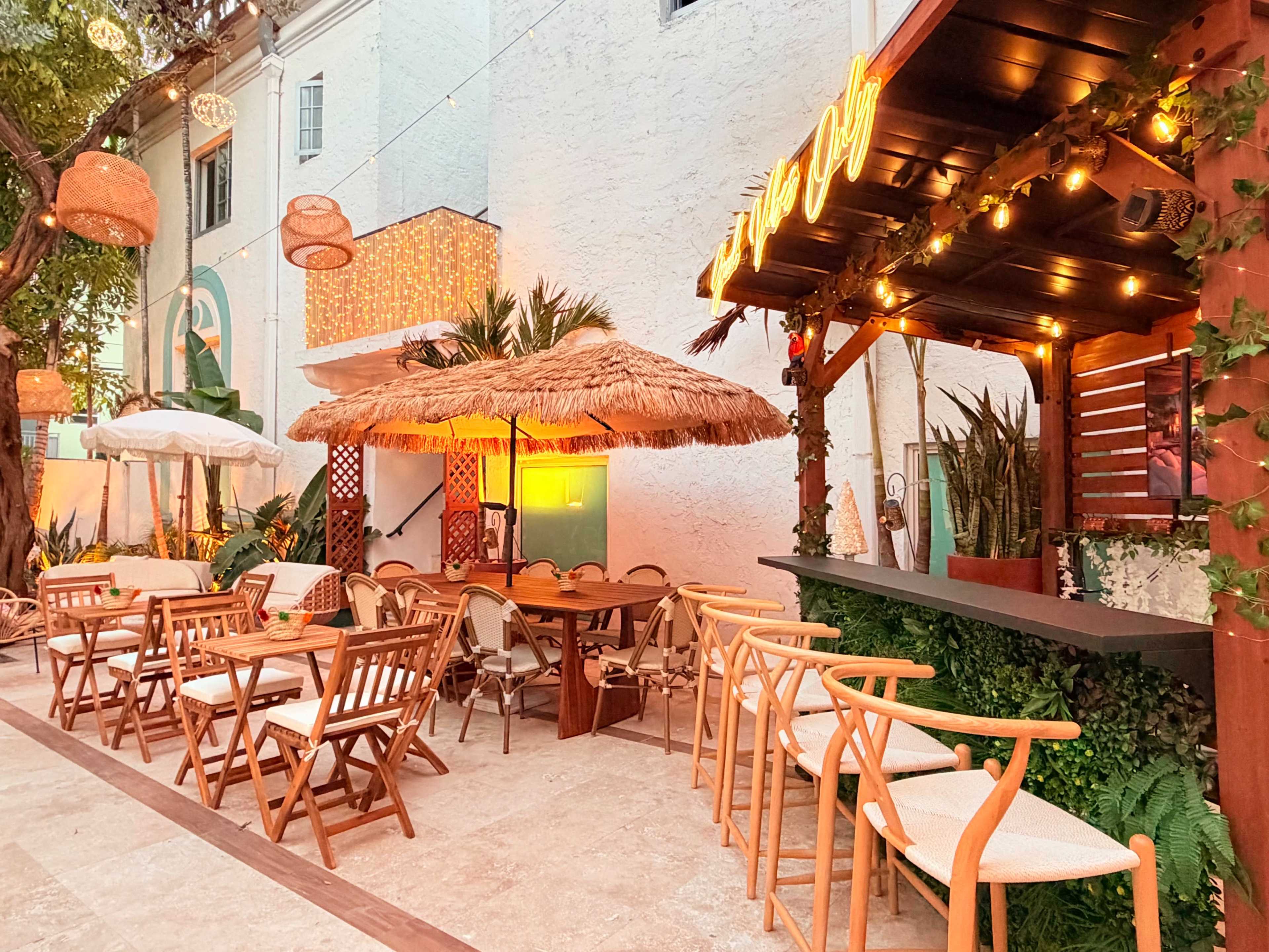 A vibrant outdoor dining area features wooden tables and chairs, a thatched roof pergola, and string lights illuminating a bar setup against a backdrop of greenery.
