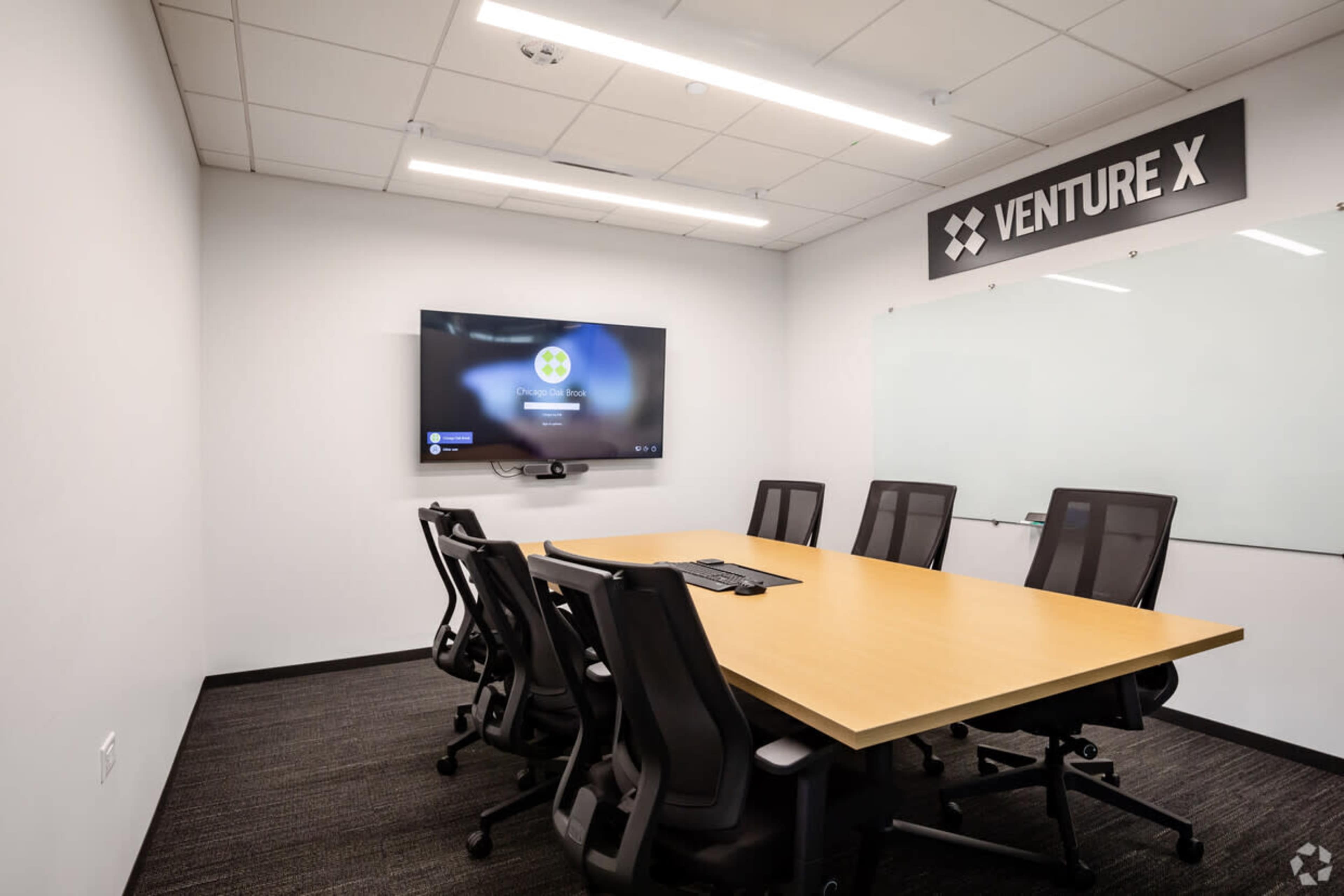 A modern conference room features a large wooden table surrounded by black office chairs, with a television mounted on the wall showing a presentation screen.