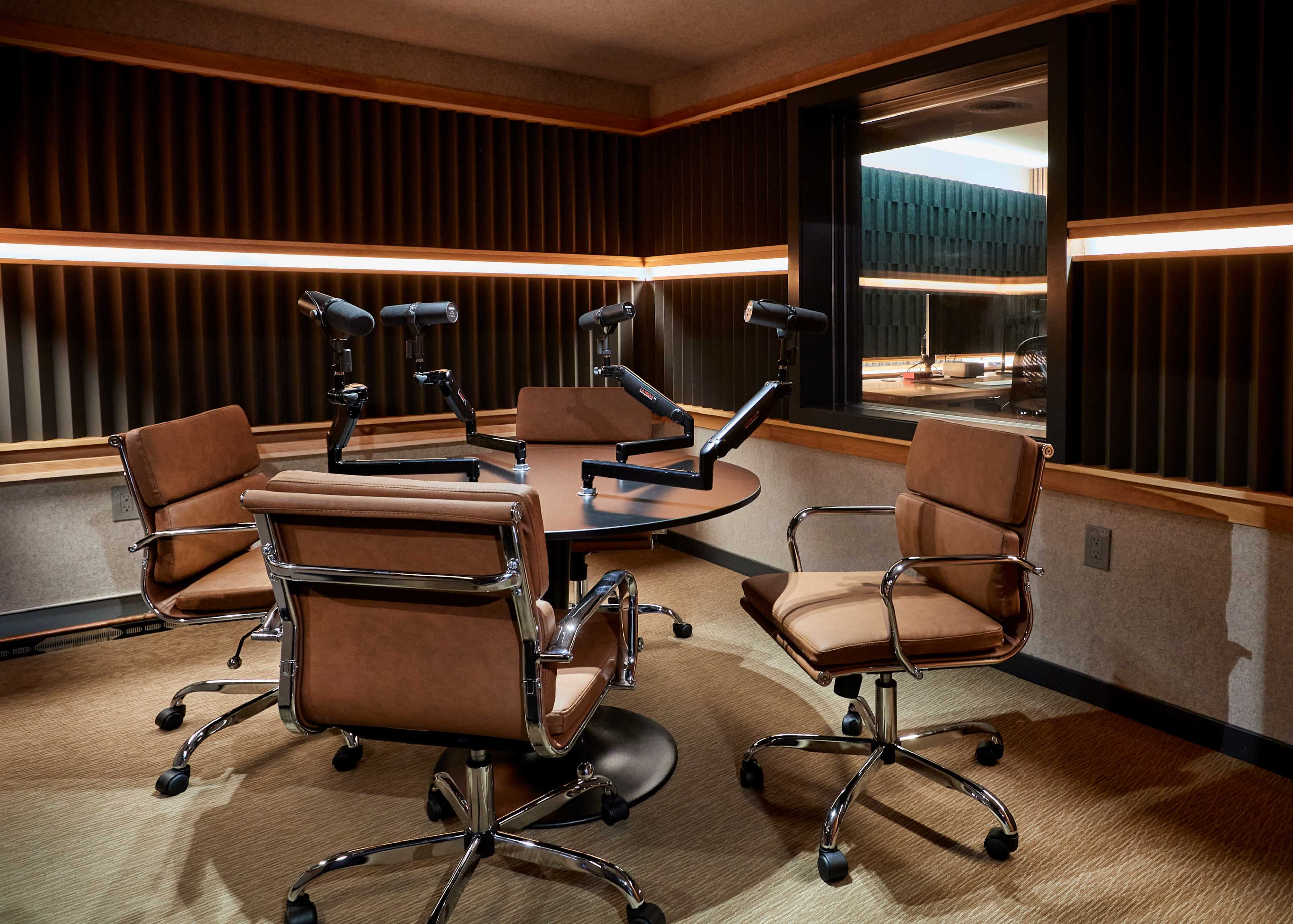 A conference room with a round wooden table surrounded by four swivel chairs, each equipped with microphone arm mounts.