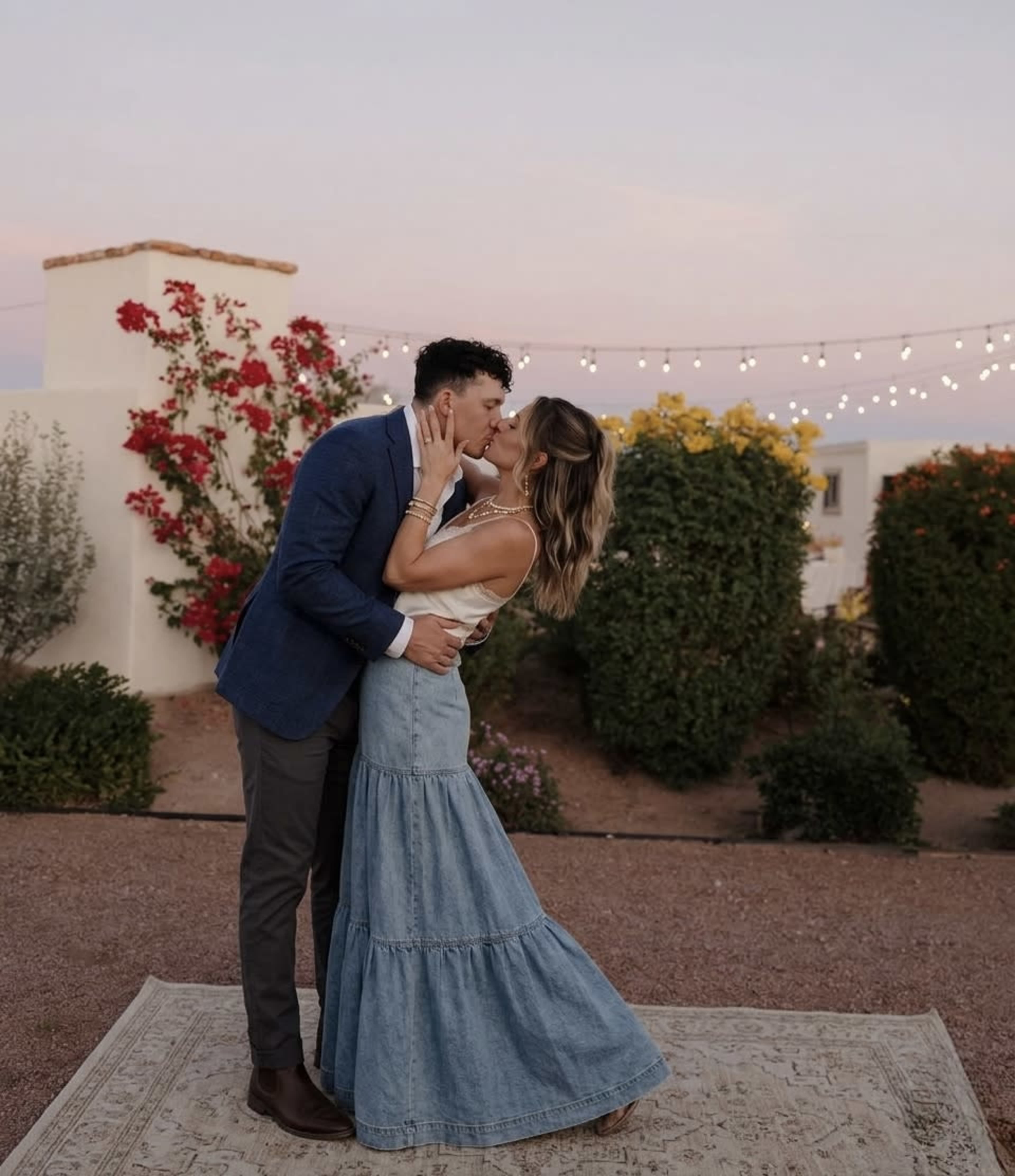 A couple kisses in a garden setting decorated with string lights and blooming flowers during twilight.