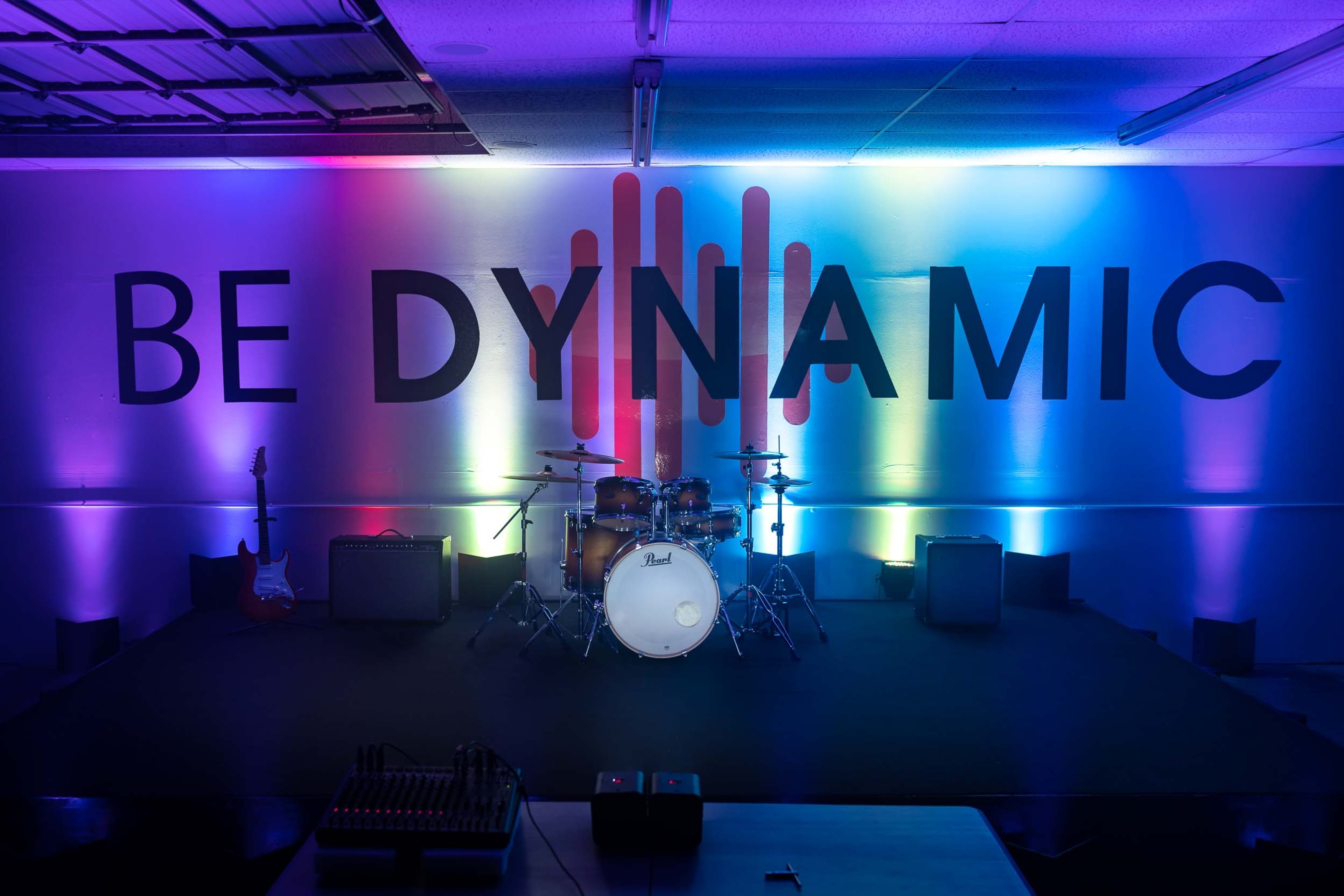 A stage setup with musical instruments in front of a brightly lit backdrop featuring the phrase "BE DYNAMIC" in large letters.